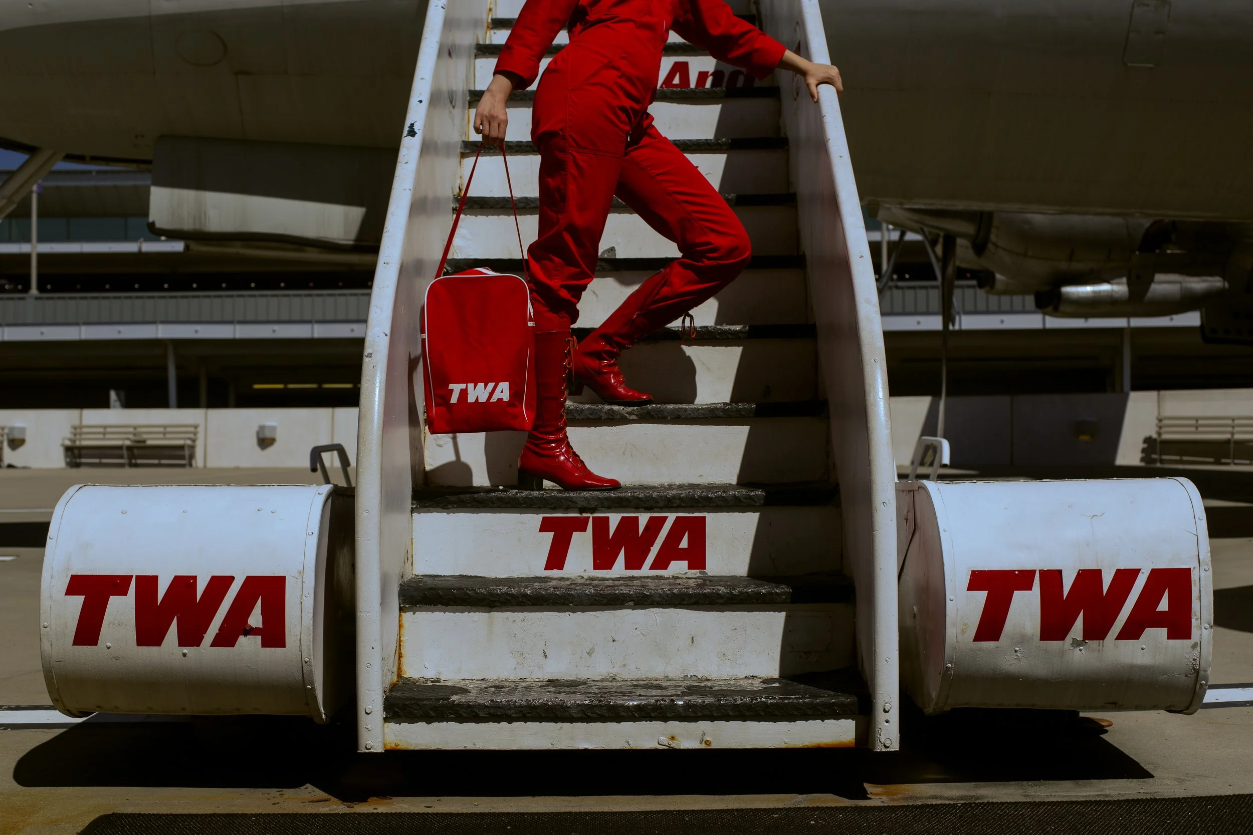 An empty airplane boarding staircase with red TWA logos, with a person dressed in red fashion standing on the stairs, holding a red bag, at an airport.