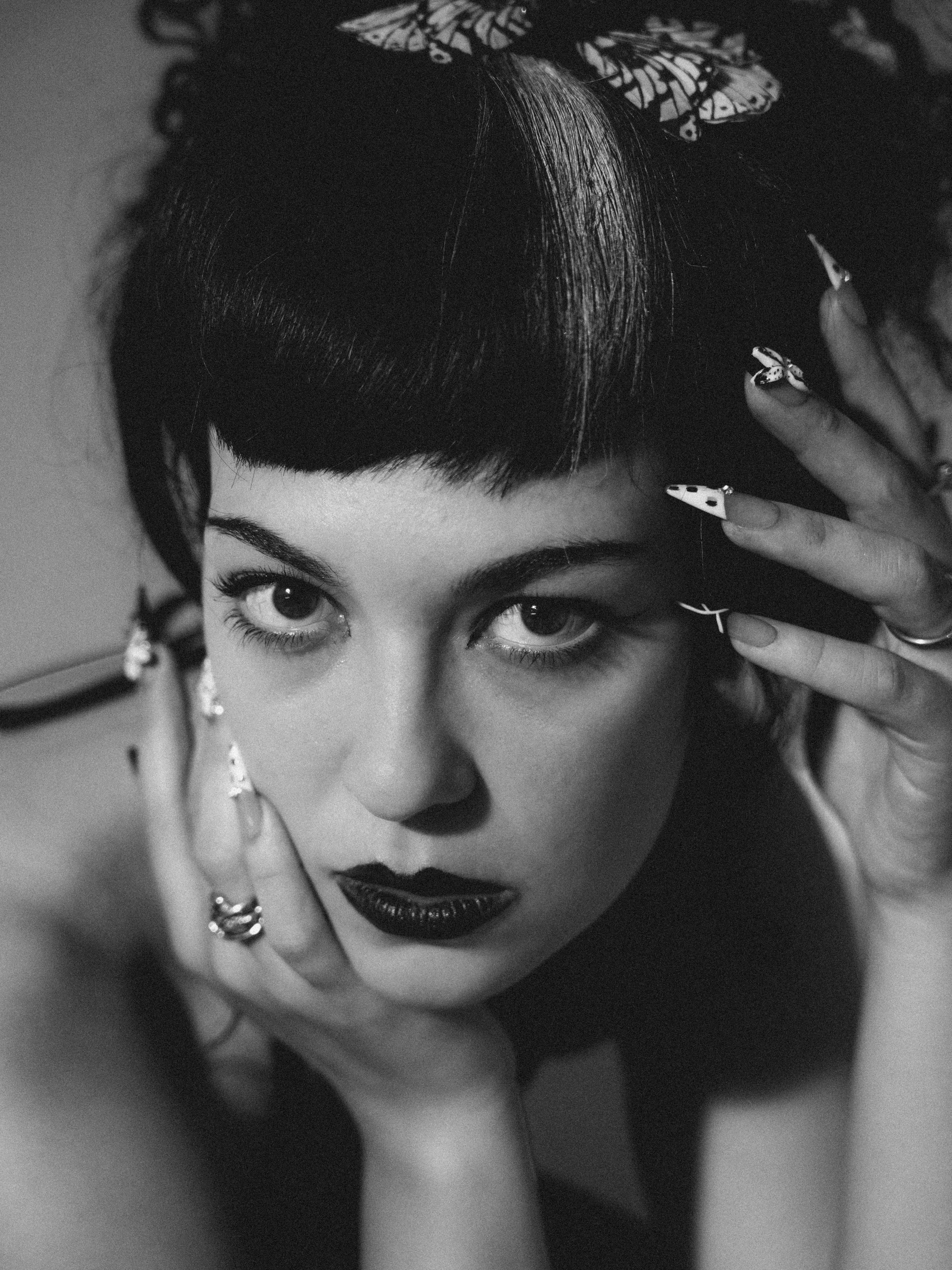 Black and white photo of a young woman with dark lipstick, dark eye makeup, and eye contact with the camera. She has dark hair with a streak of light color and is wearing butterfly hair clips and jewelry.