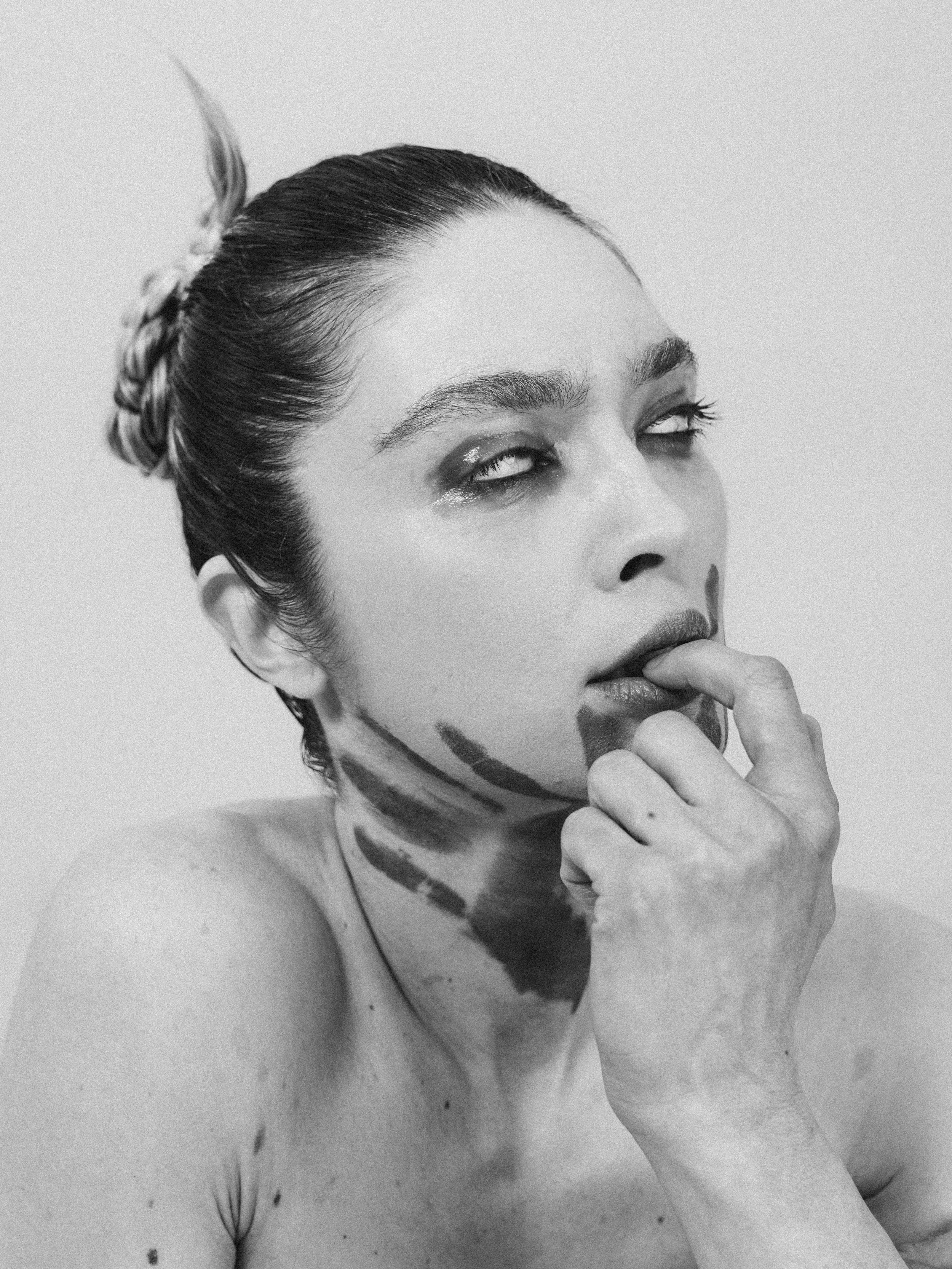 A black and white photo of a woman with her hair in a bun and face painted with geometric shapes, holding her chin with her hand.