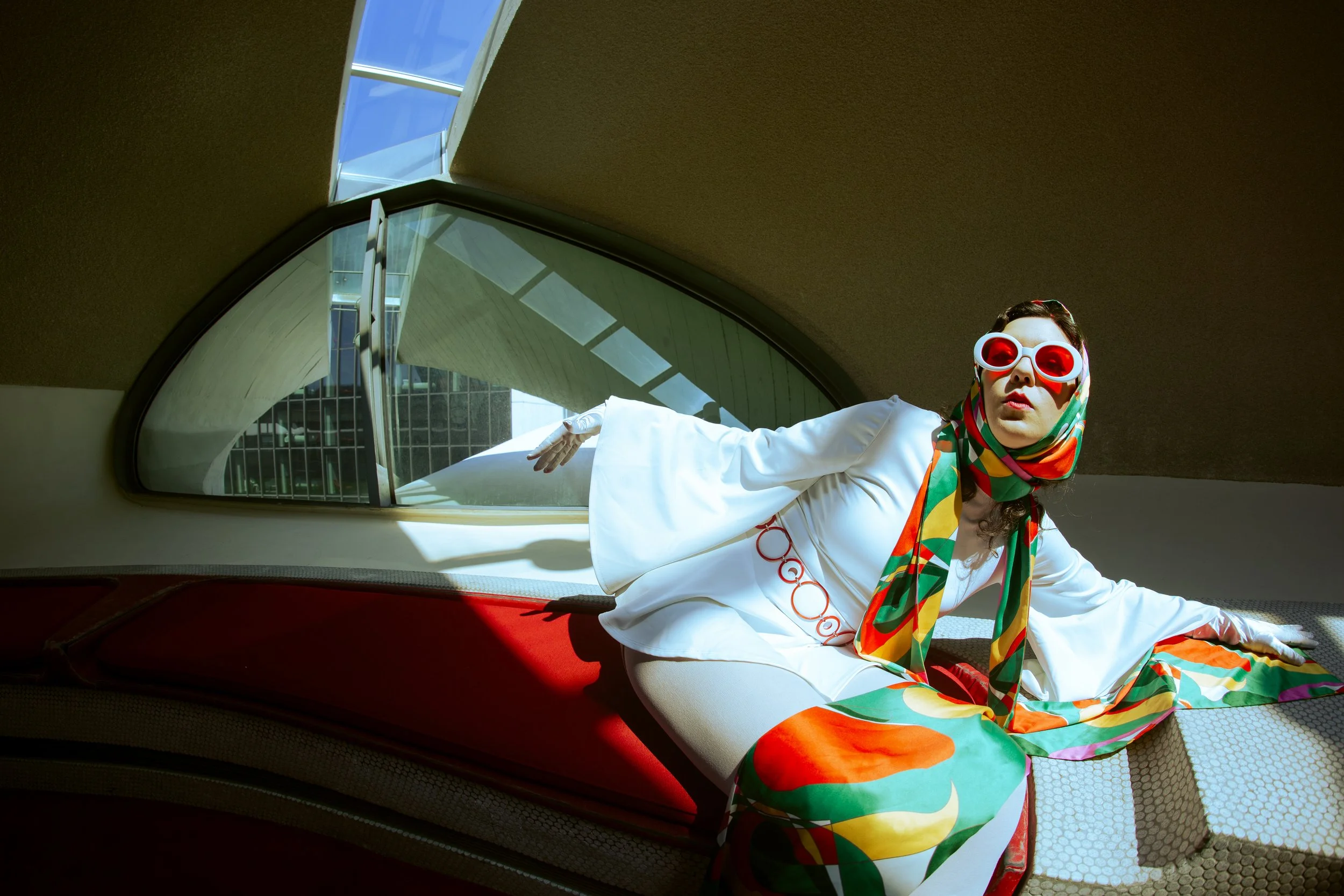 Woman wearing white coat, colorful scarf, and large red sunglasses lying inside a stylish building with modern architecture and large windows.