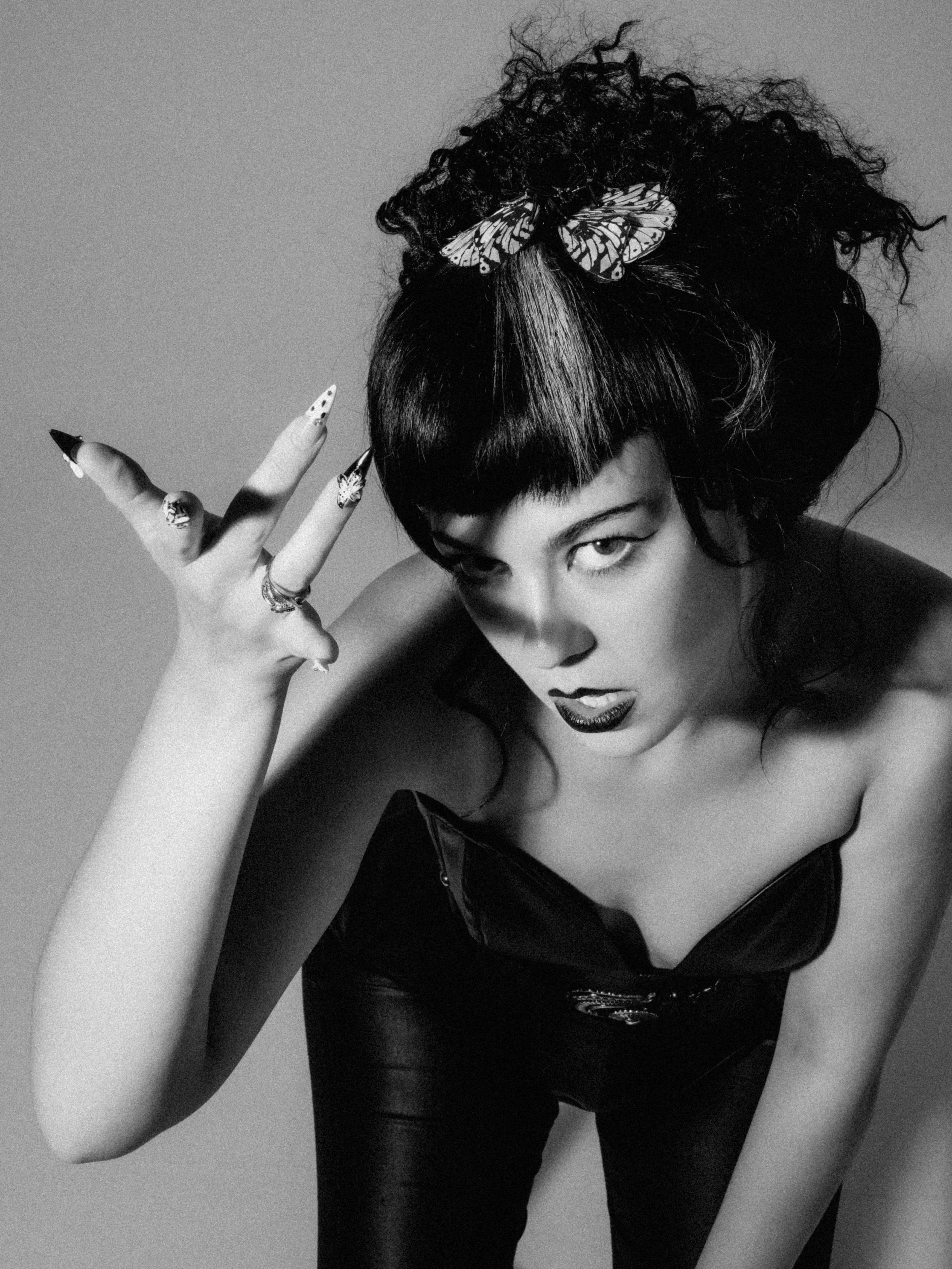 A woman with dark curly hair and a streak of lighter hair, wearing dark lipstick and eye makeup, holding up her hand with elaborately decorated fingers and rings, posing with a serious expression in a black leather outfit with a zipper.