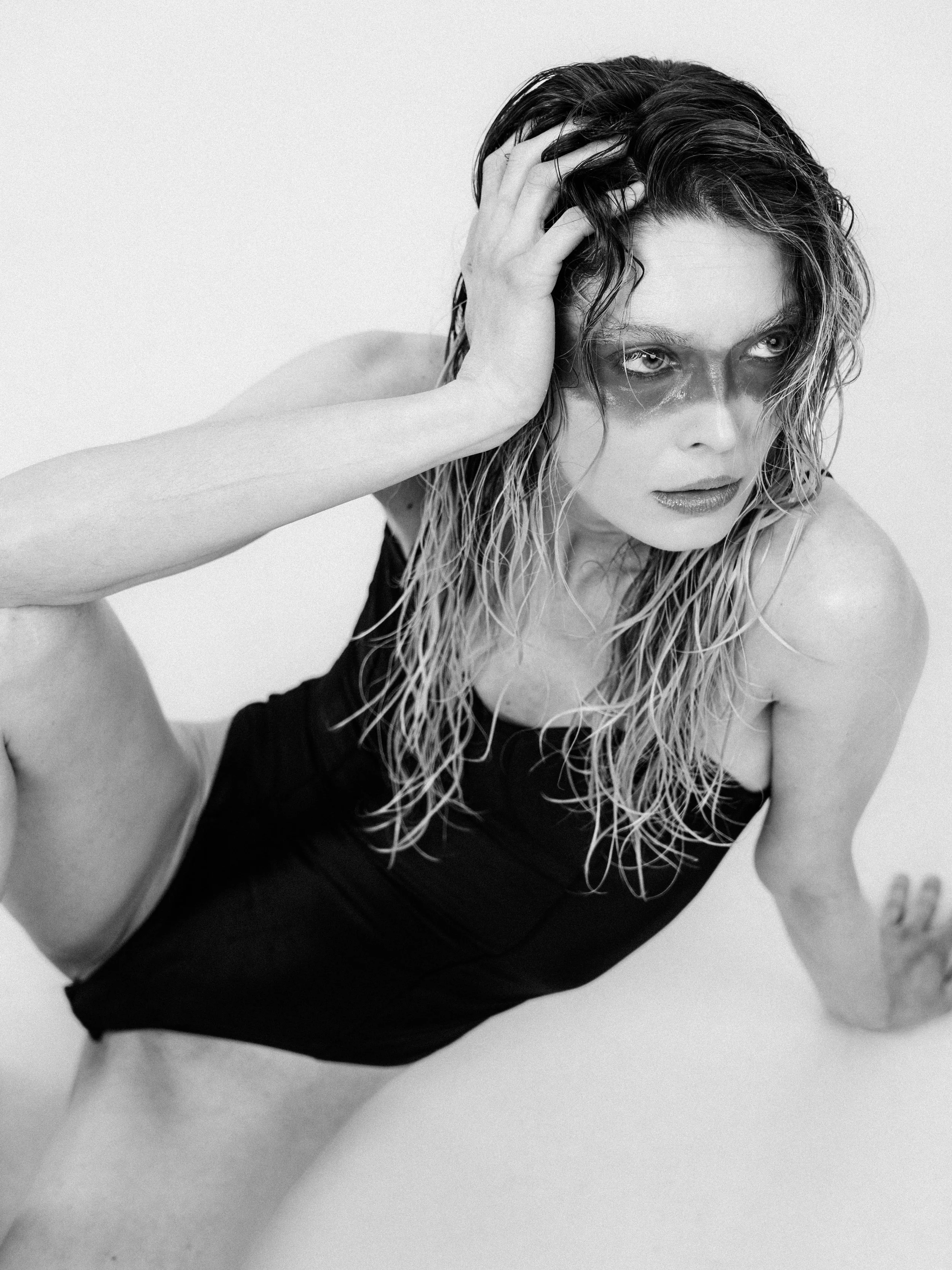 Black and white photograph of a woman with wet, curly hair, posing with her hand on her head, wearing a sleeveless black top, against a plain background.