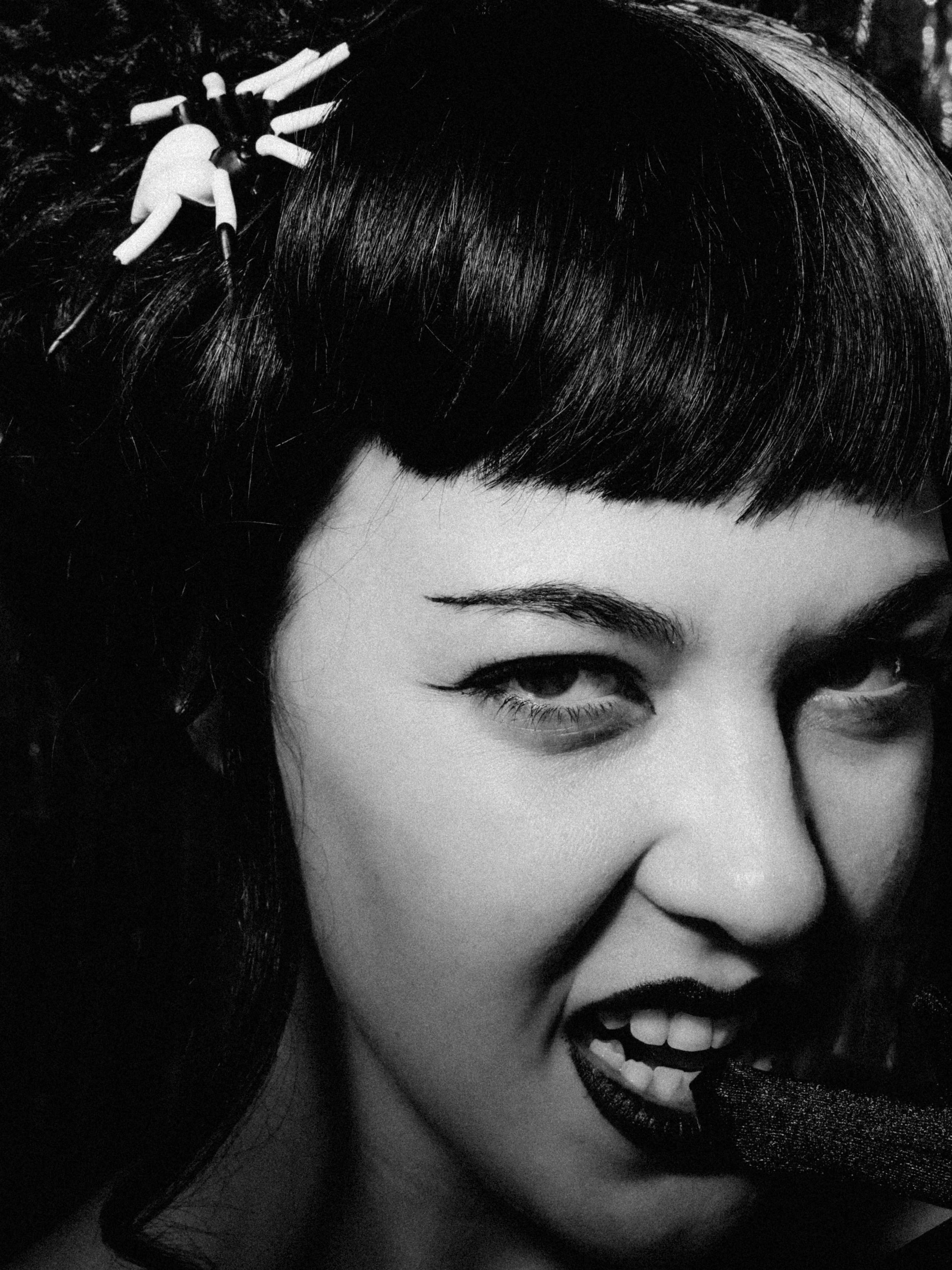 Black and white photograph of a woman with dark, shiny hair styled with a bow-shaped hair accessory, smiling and showing her teeth with dark lipstick, eye makeup, and visible eyebrows.