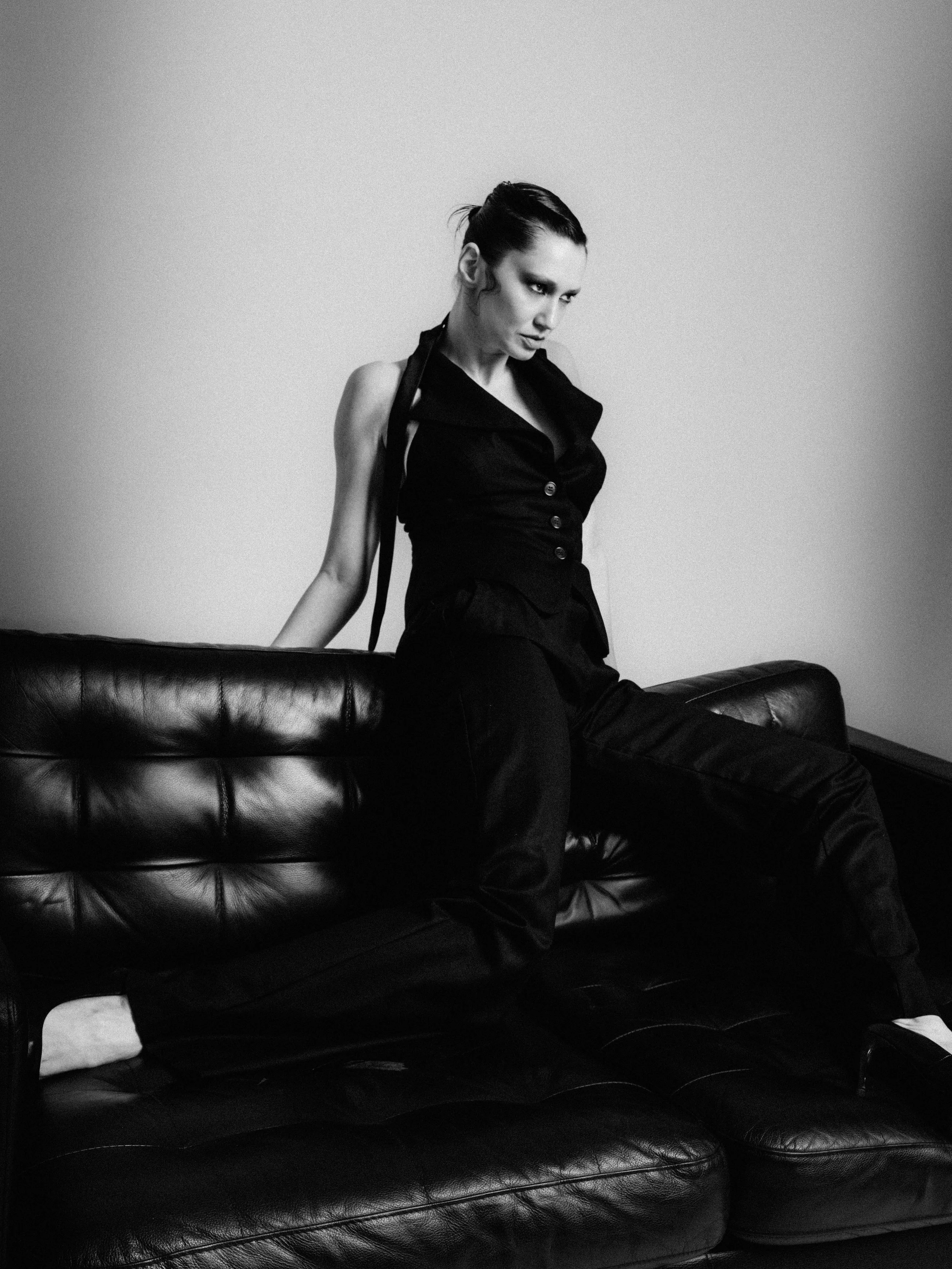 A black-and-white photo of a woman with short hair, wearing a sleeveless dark blouse with buttons, sitting on a leather couch with her left arm resting on its back, looking contemplative.