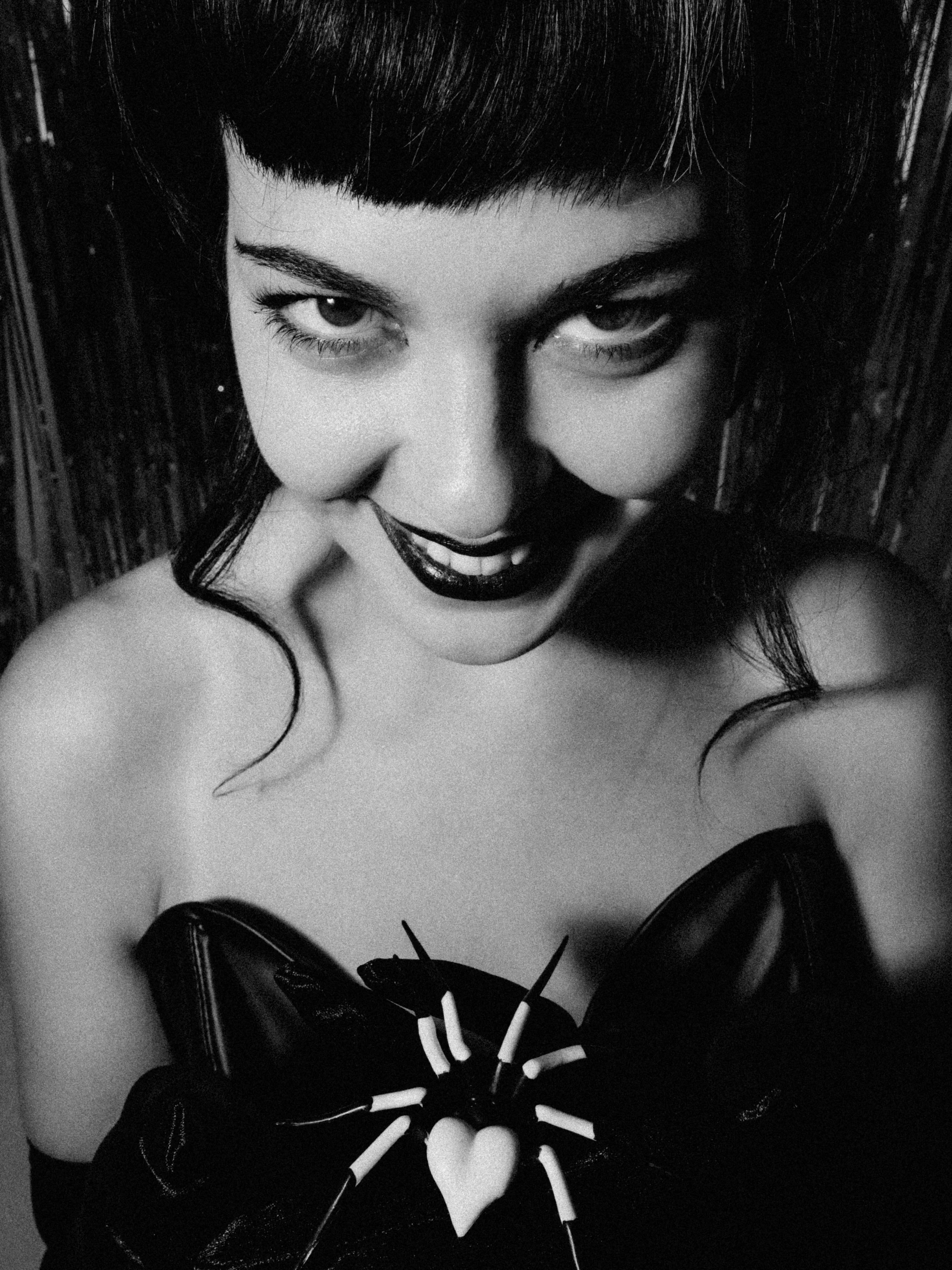 Black and white photo of a woman with dark hair, wearing dark lipstick and gloves, holding a heart-shaped lollipop.