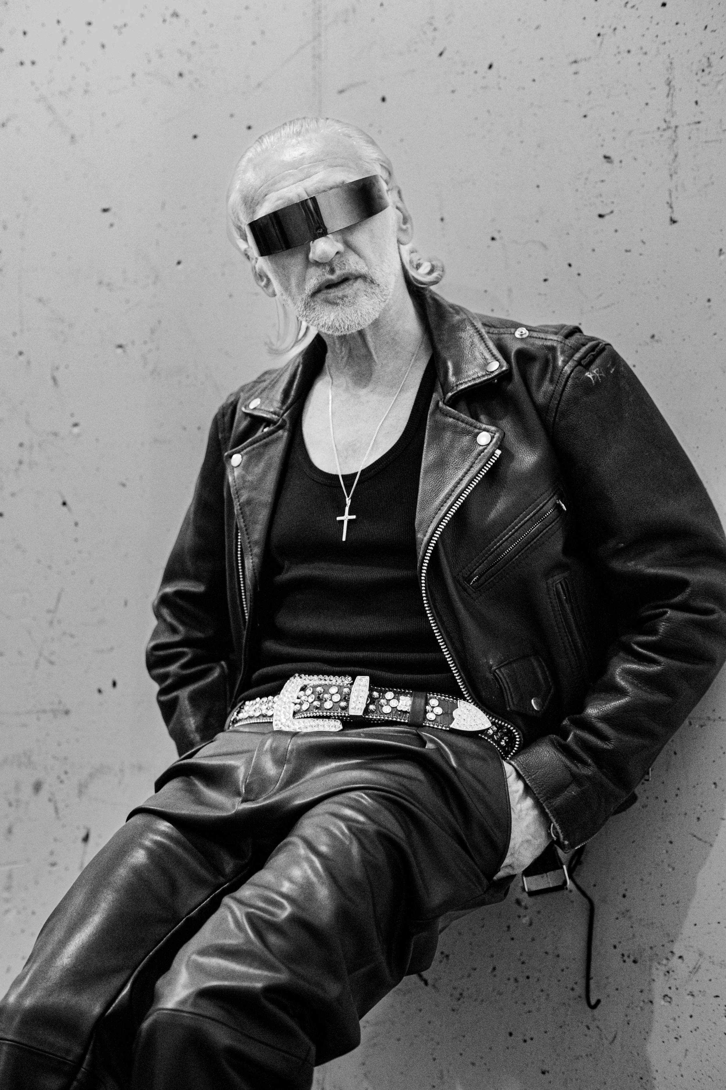 Black and white photo of a man with long hair and beard, wearing sunglasses, leather jacket, necklace with cross, and ornate belt, sitting against a plain textured wall