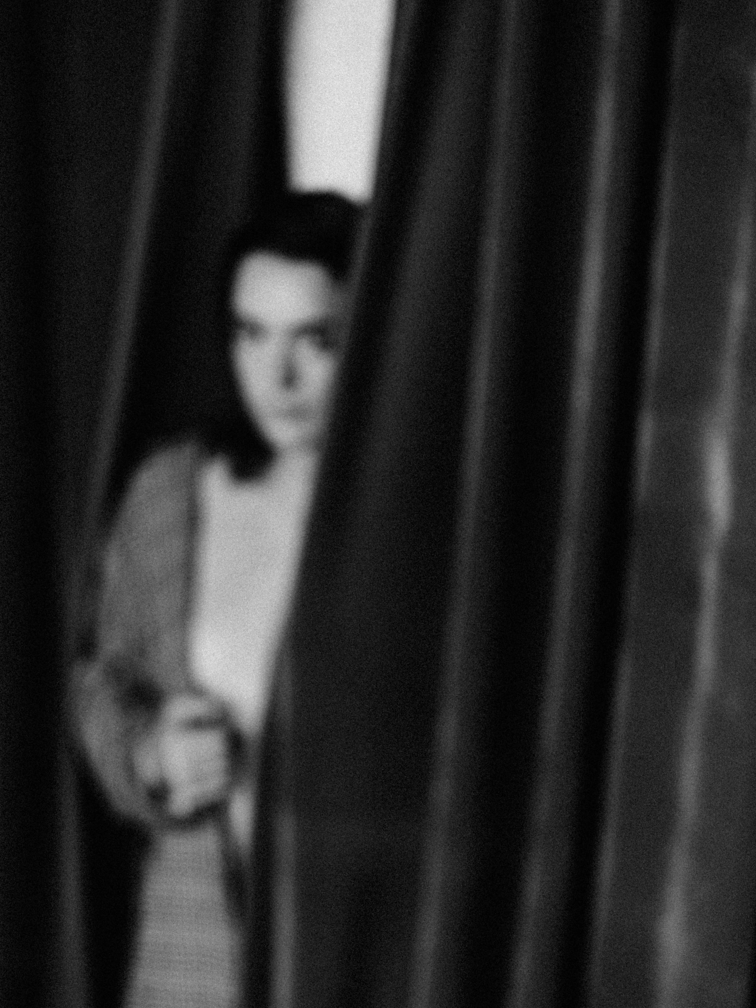 Blurred black and white photograph of a person's face partially obscured by vertical blinds or curtains.
