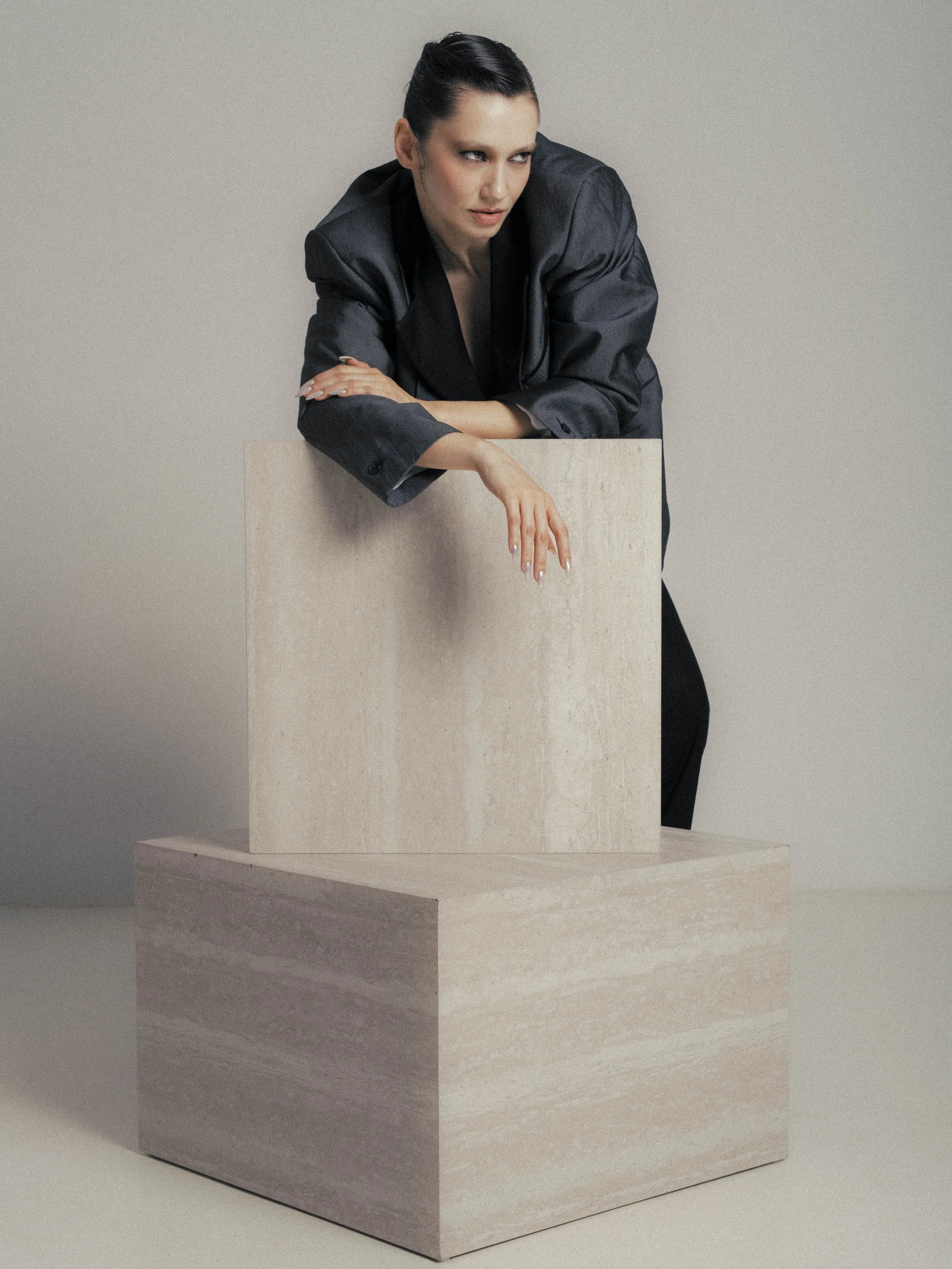 A woman with dark hair styled in a sleek bun wearing a black satin blazer, leaning on a large, curved beige sculpture, with her right arm resting on top and her left hand hanging down, against a plain light-colored background.