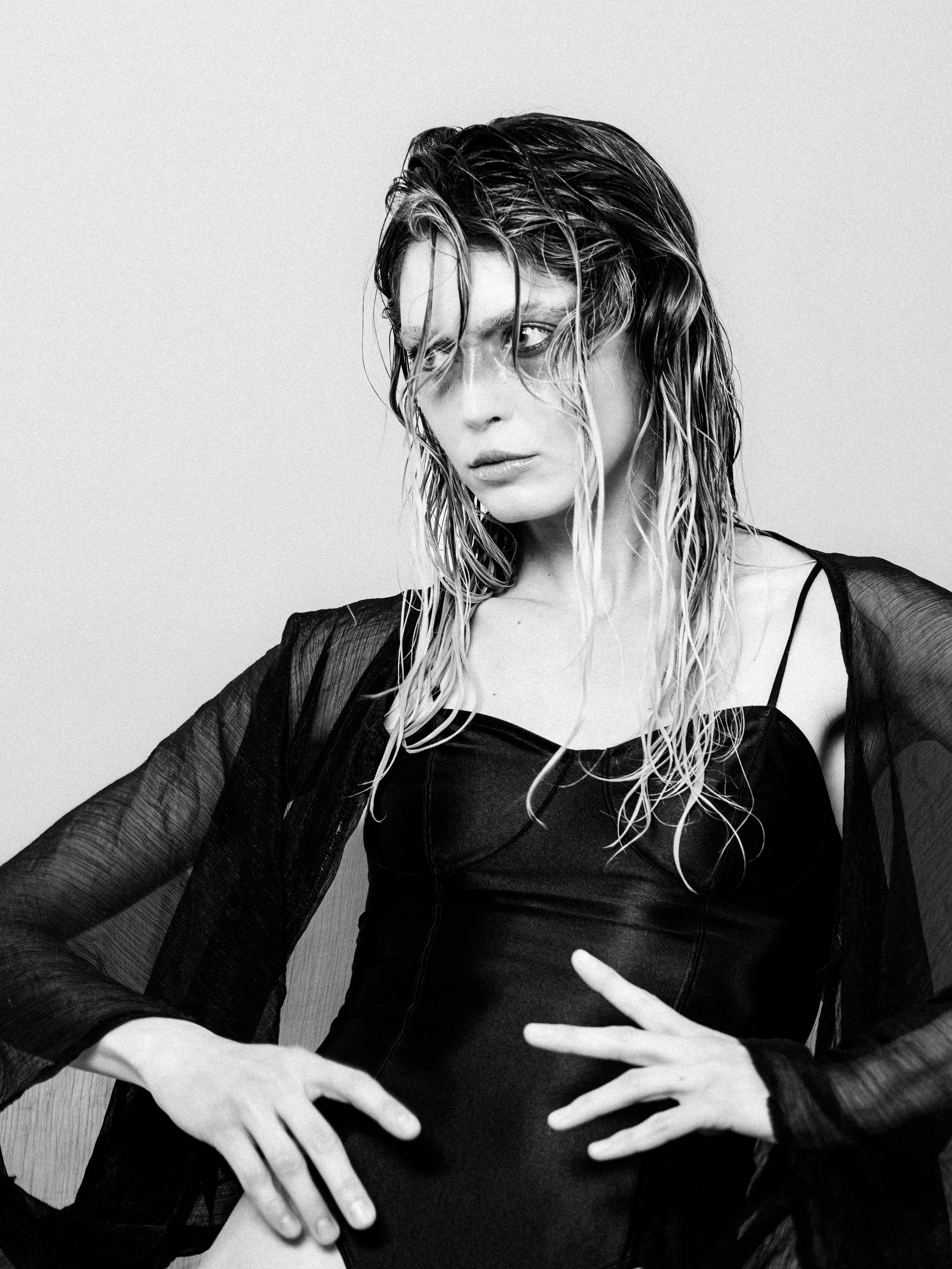 Black and white photo of a woman with wet hair, wearing a black top with sheer sleeves, posing with her hands on her waist and slightly tilting her head.