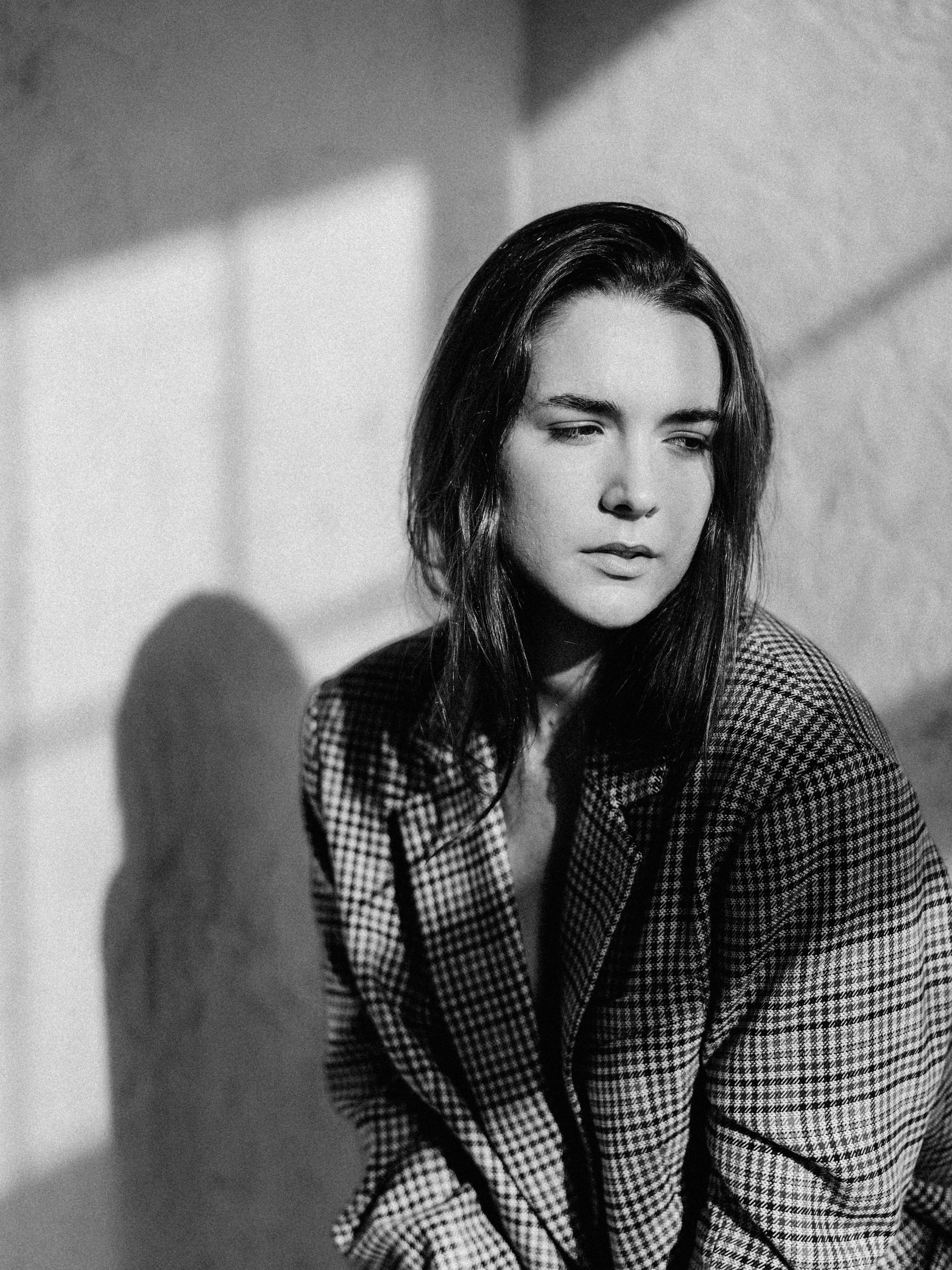 A black and white photograph of a young woman with long dark hair and closed eyes, dressed in a checkered blazer, facing slightly downward, with soft shadows cast on the wall behind her.
