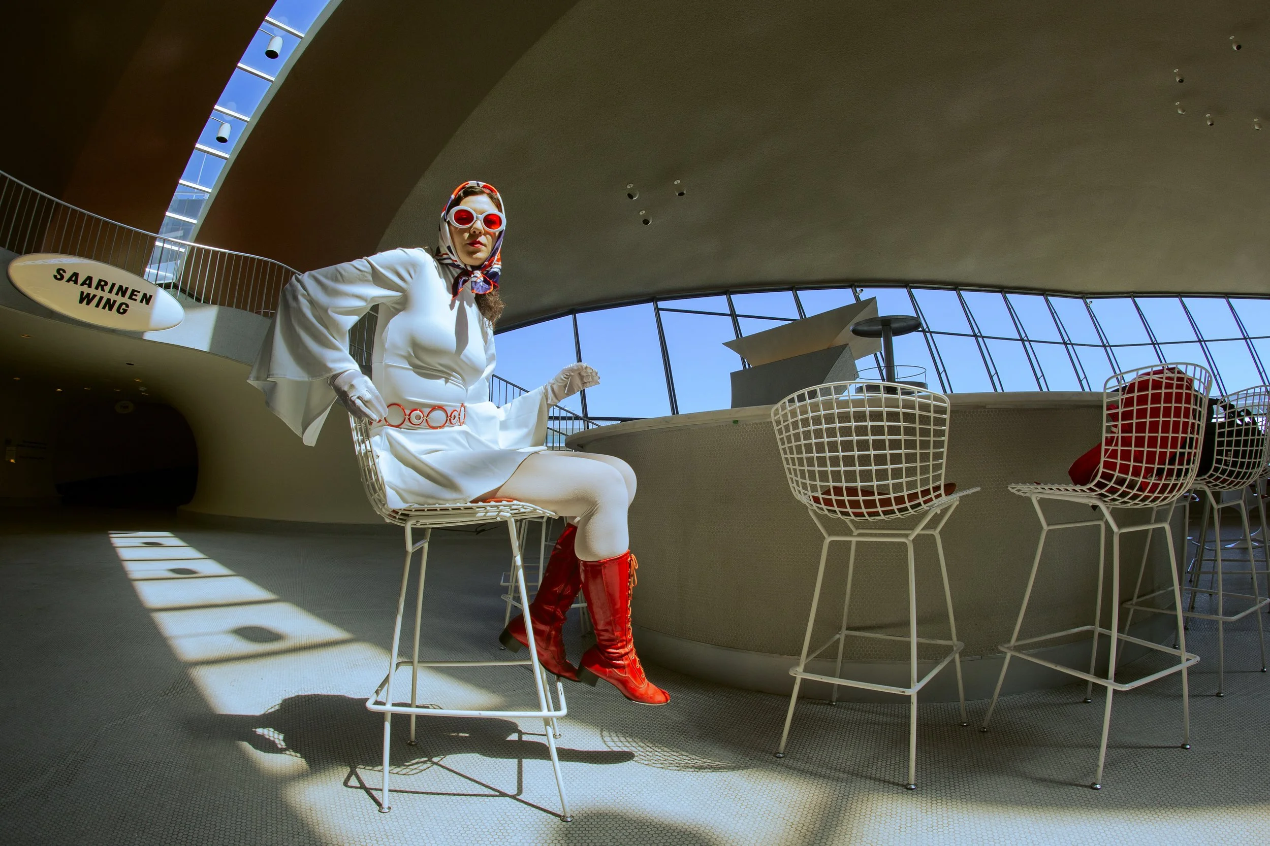 Woman in vintage 60s style outfit with red boots, white dress, red accessories, and large sunglasses sitting on a white wire chair in a modern building with large glass windows and curved architecture.