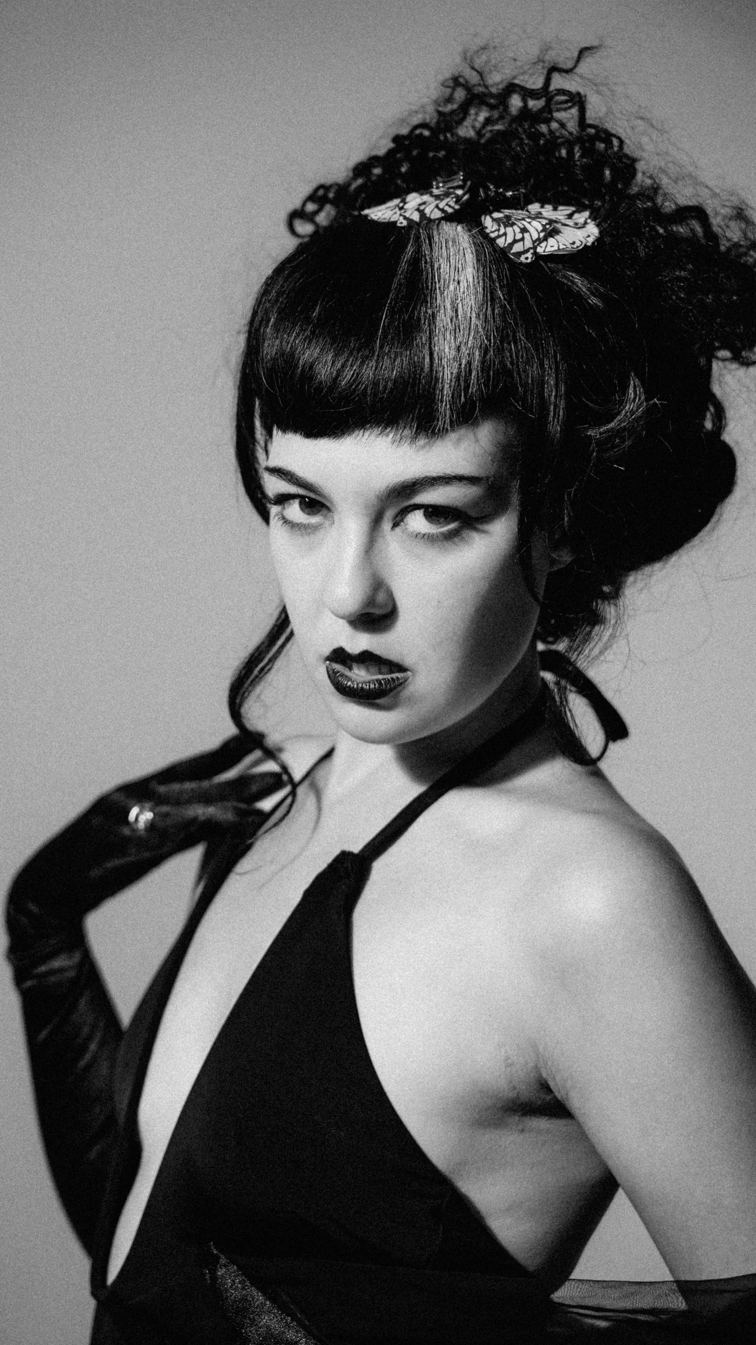 Black and white photograph of a woman wearing a leather glove, with dark lipstick, dark eye makeup, wearing a butterfly hair clip, and a revealing black outfit with straps.