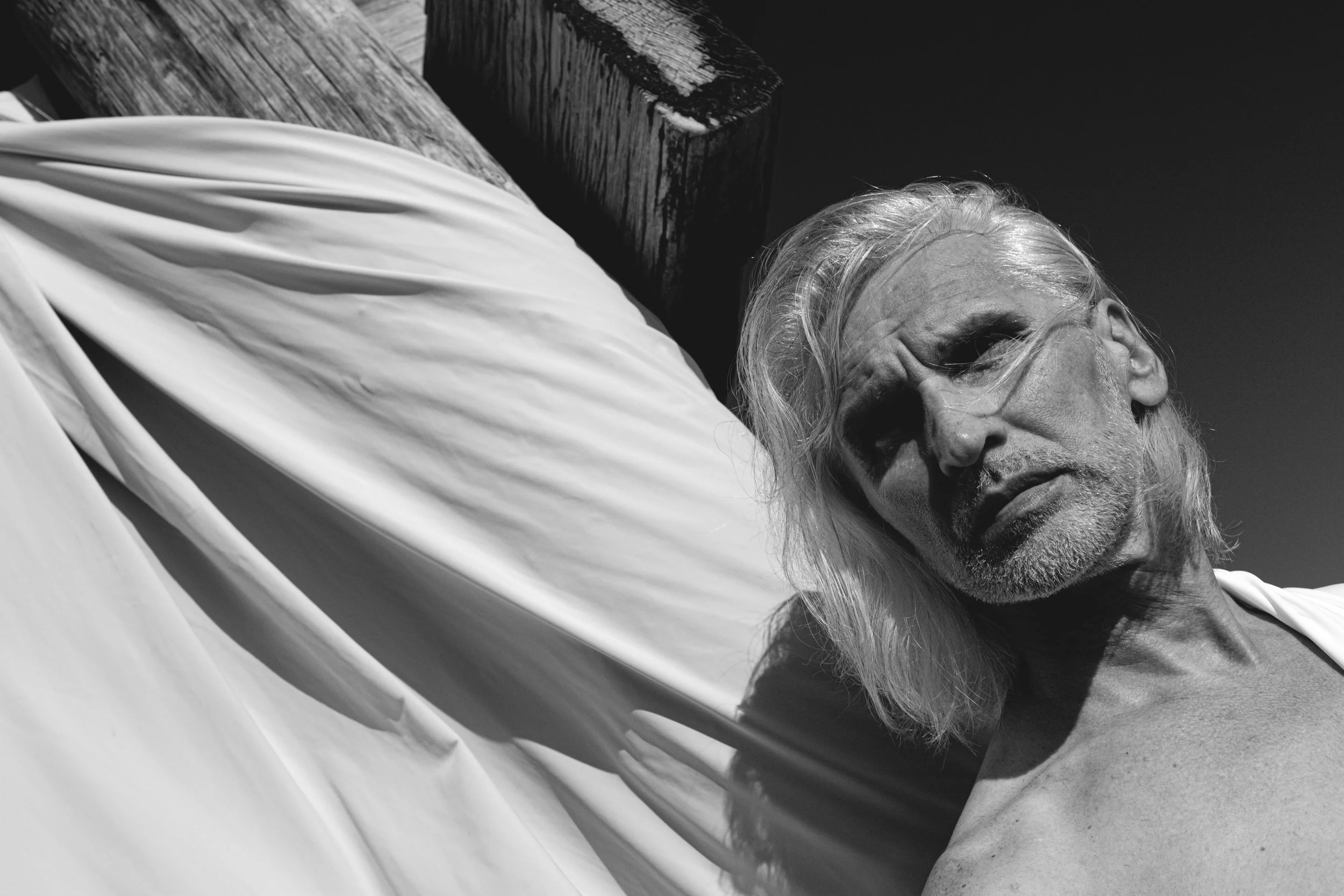 A black and white photo of a man with long hair and a beard lying on his side on a wooden surface, with a cloth partially covering him.