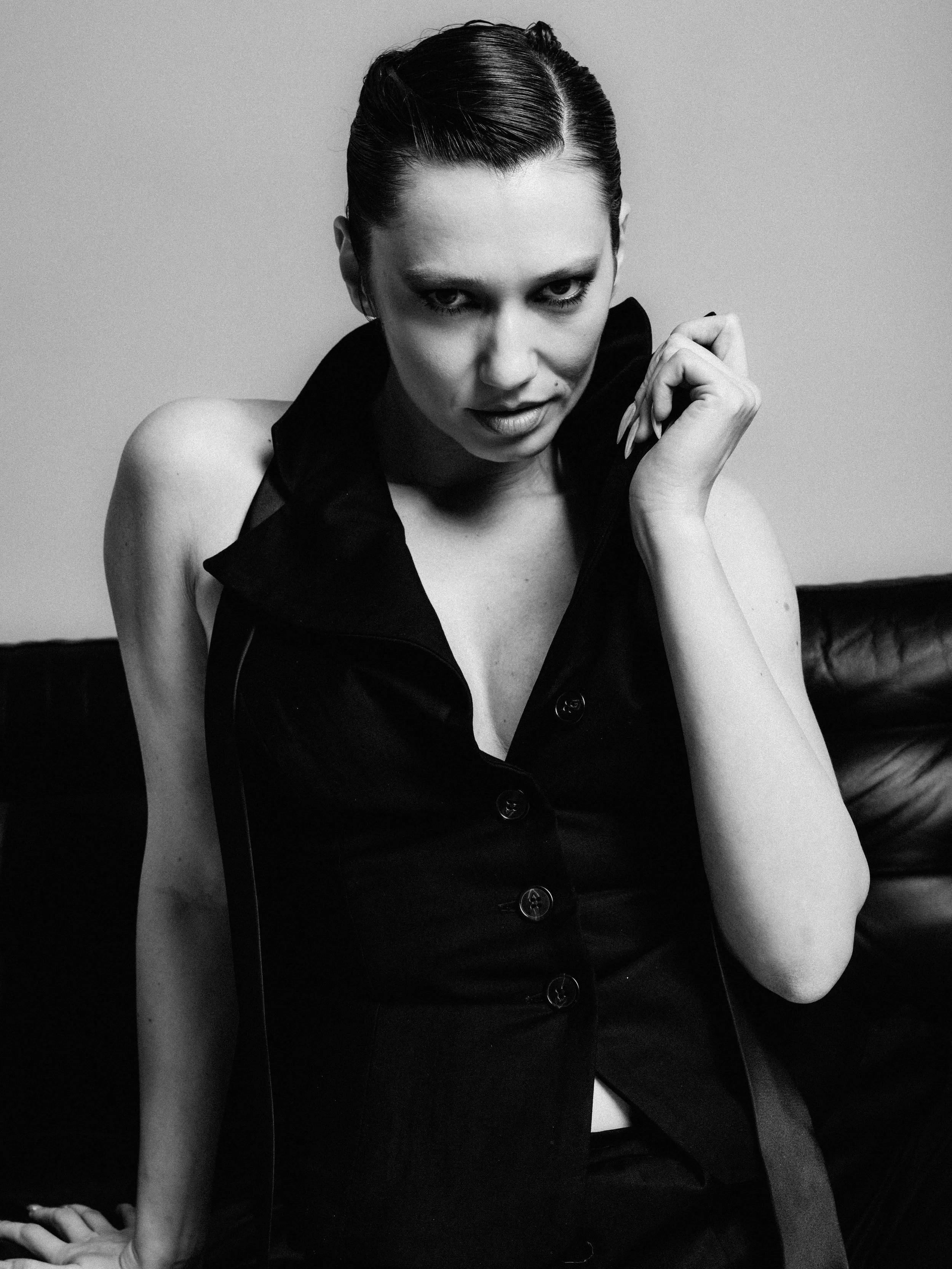 A black and white photograph of a woman with slicked-back hair, wearing a sleeveless black dress with buttons down the front, sitting on a sofa with her left arm resting on the ground and her right hand near her face. She is looking directly at the c