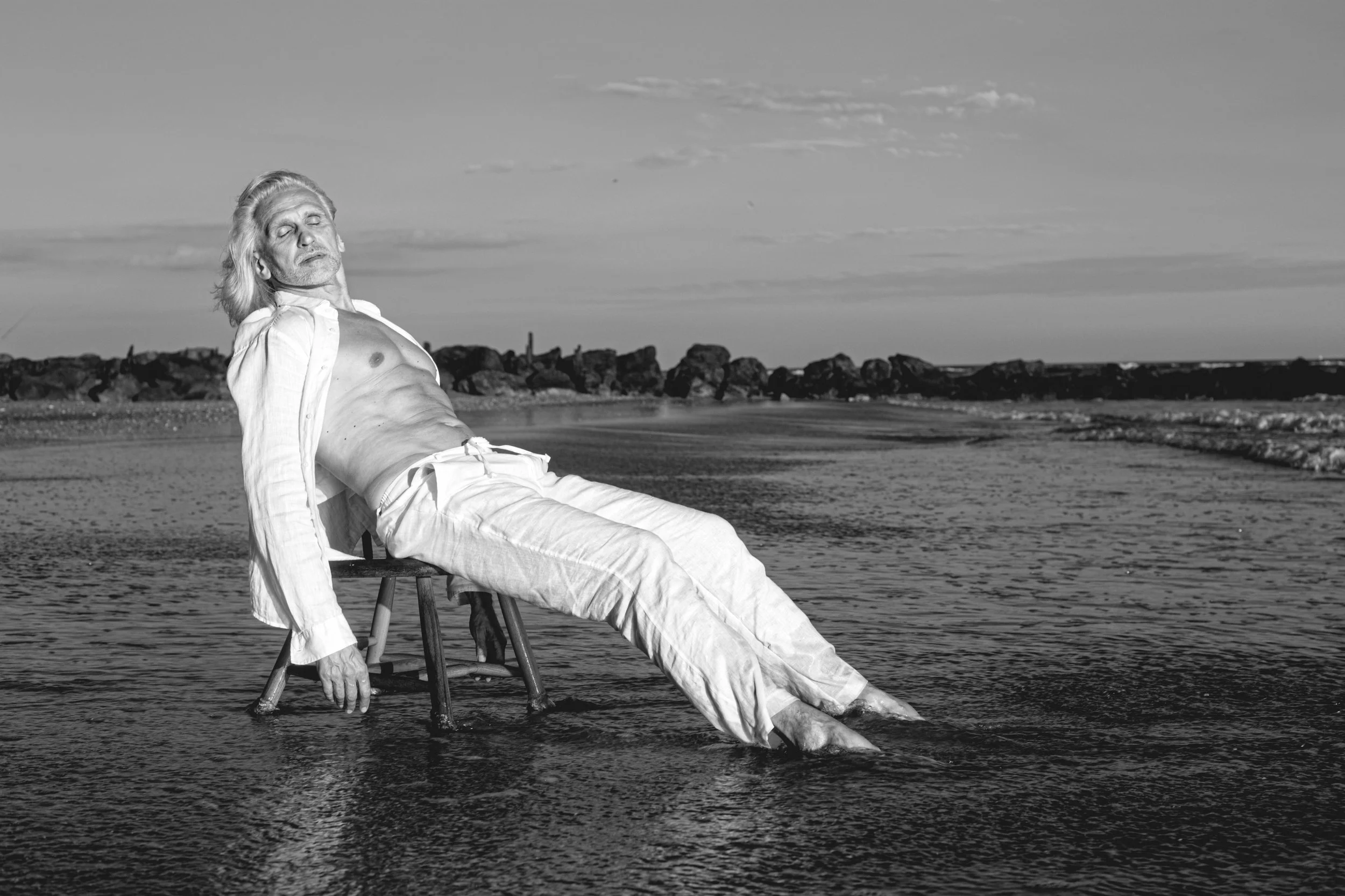 A black and white photo of an older man with long hair and a beard, sitting on a stool in shallow water at the beach, with his eyes closed and head turned slightly upward, wearing an open shirt and white pants.