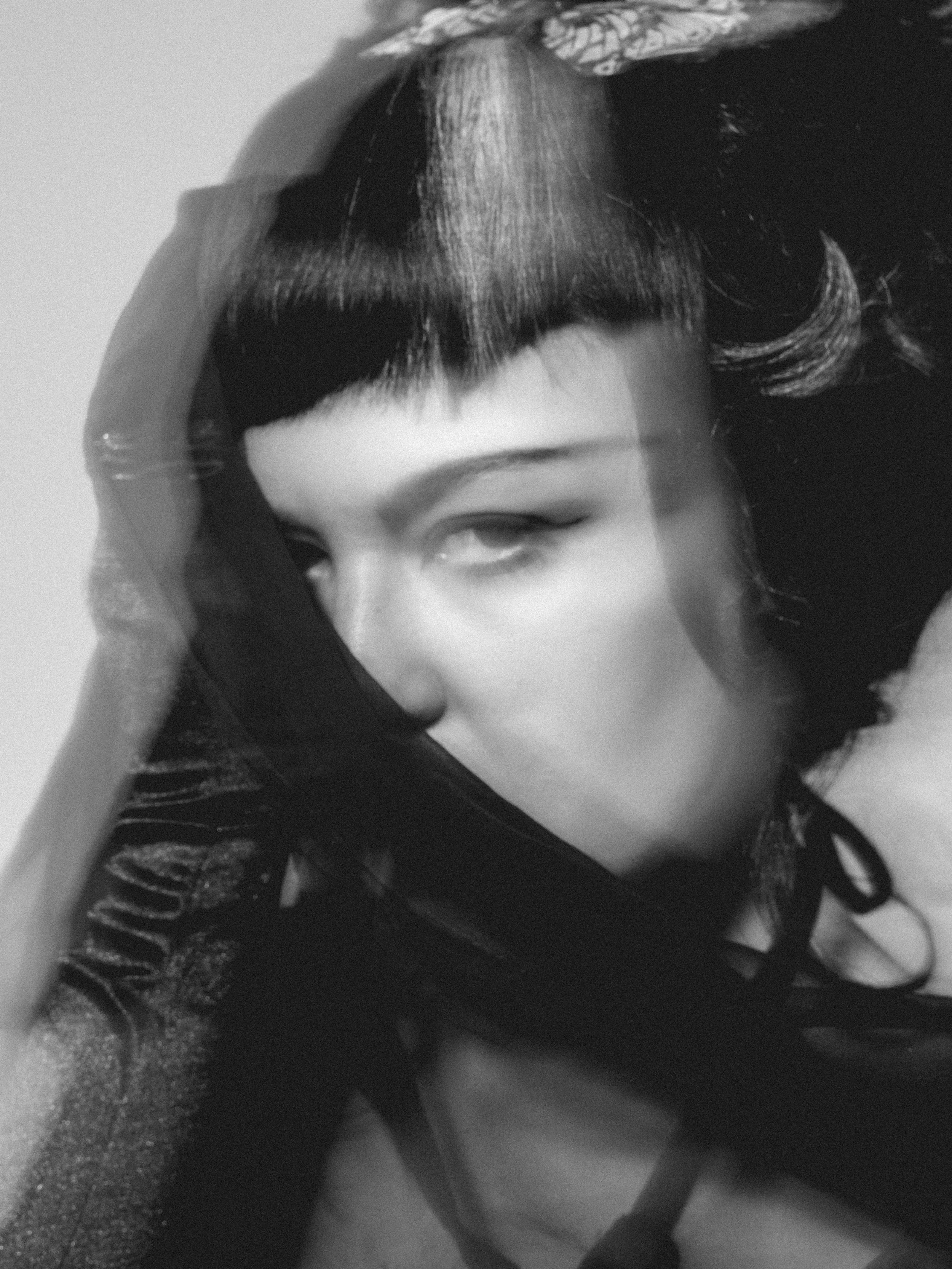A black and white close-up of a young woman with short, styled hair, wearing a dark top, with her eyes half-closed, lying down on a textured surface and holding a piece of cloth or fabric near her head.