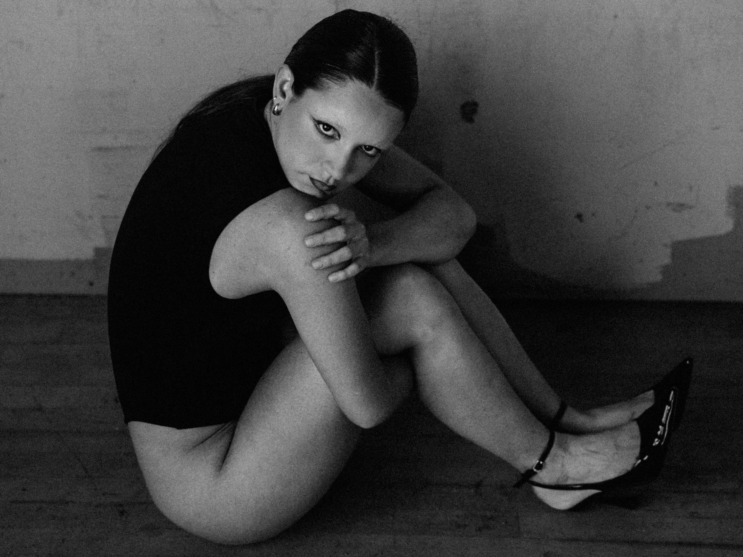A woman with dark hair styled back, wearing earrings, sitting with her arms around her legs, looking directly at the camera in a black-and-white photograph.