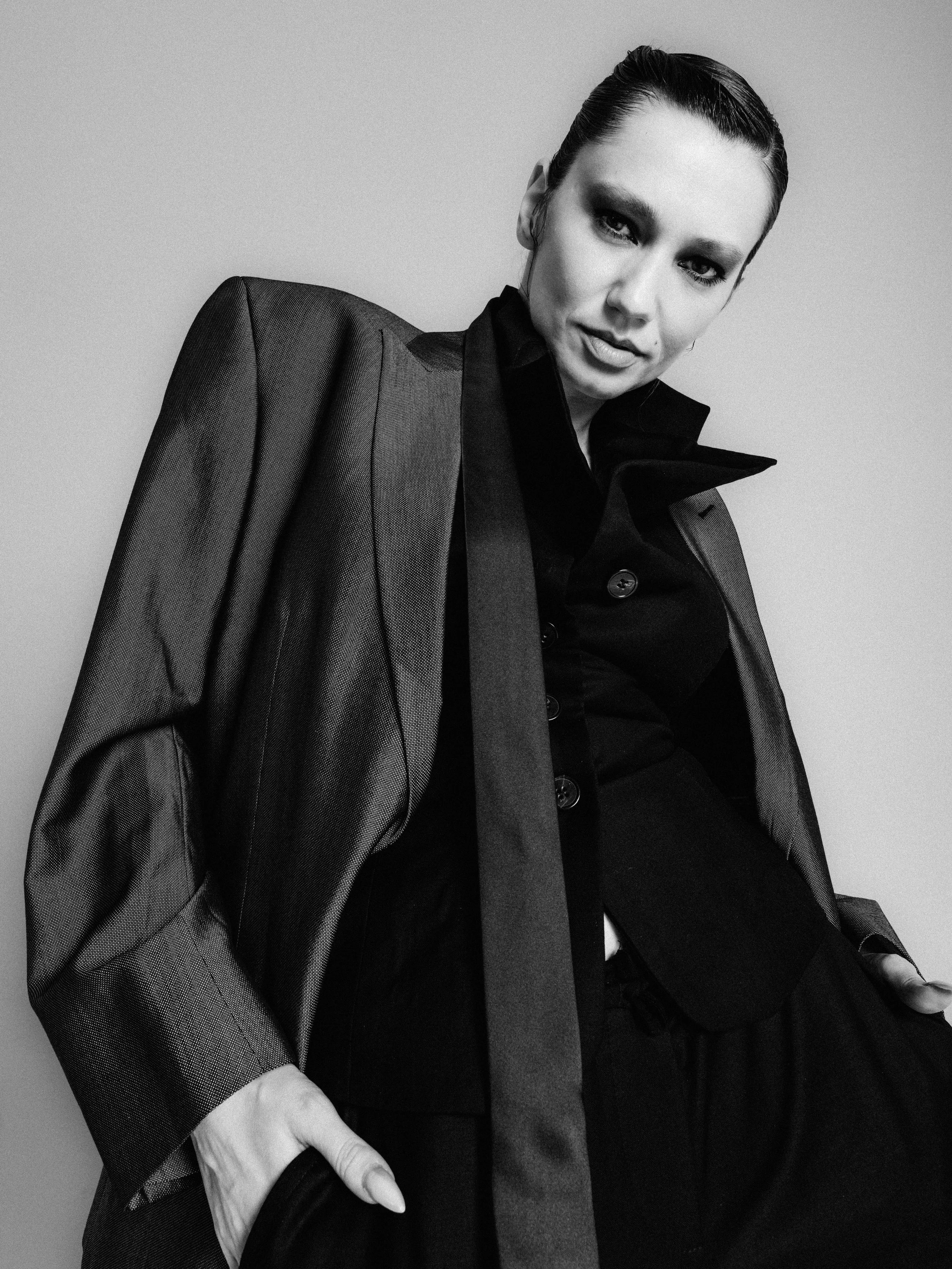 A black and white photo of a woman with slicked-back hair wearing a dark blazer draped over her shoulders. She has a confident expression and is leaning slightly, with one hand holding her purse and the other resting on her lap.