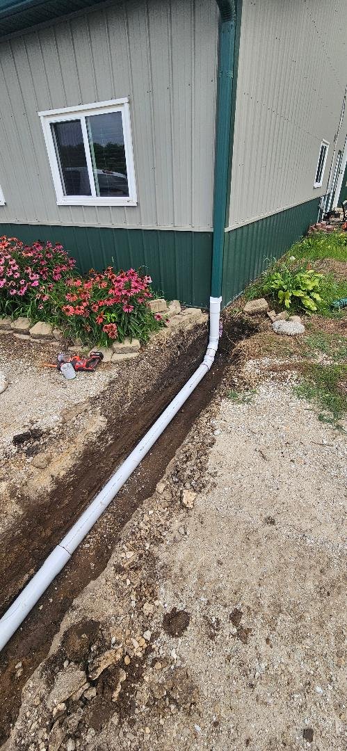 A white PVC pipe installed from the ground along the side of a house with beige siding and green trim, appearing to be part of a drainage or plumbing system, with construction dirt and a garden with pink flowers nearby.