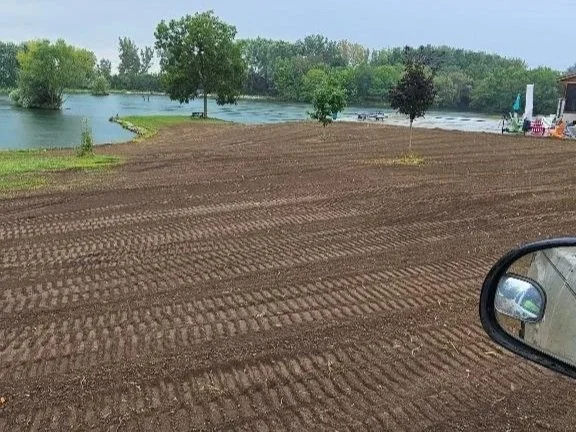 A recently tilled soil area near a lake with trees, a house, and a side mirror visible in the foreground.