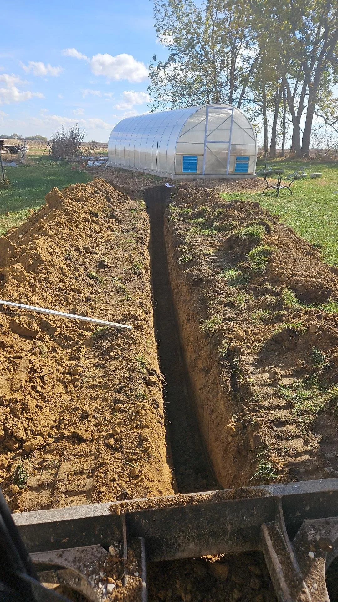 A deep trench dug into the ground with a garden bed and a greenhouse in the background. The sky is clear with a few clouds.