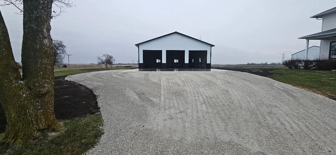 A gravel driveway leading up to a white barn with black doors, beside a house on the right, in a rural landscape with trees and an overcast sky.