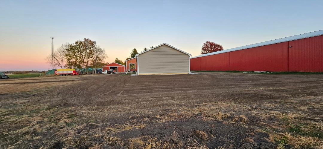 Wide view of a farm or agricultural area with buildings, trees, and a large red barn or storage building, under a sunset sky.