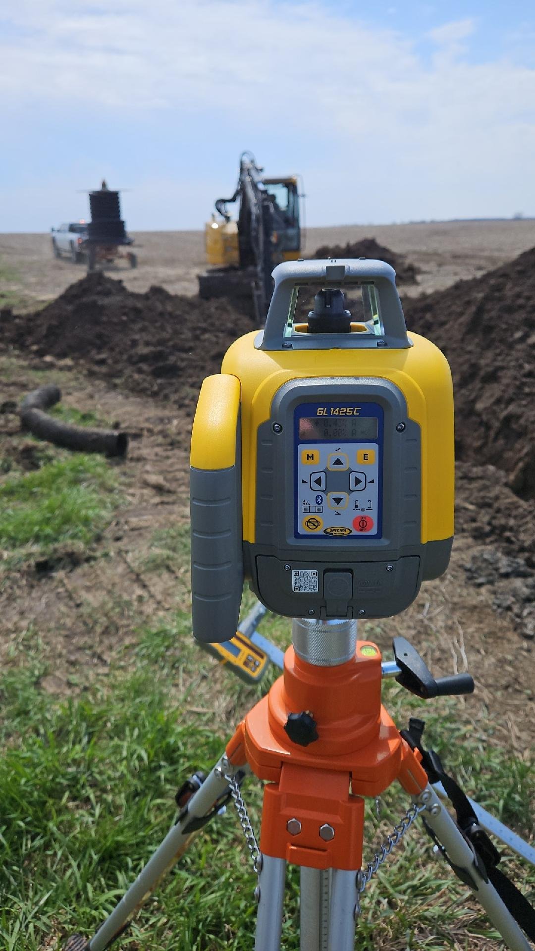 Surveying equipment set up on a tripod in an open field with a backhoe digging a trench in the background.