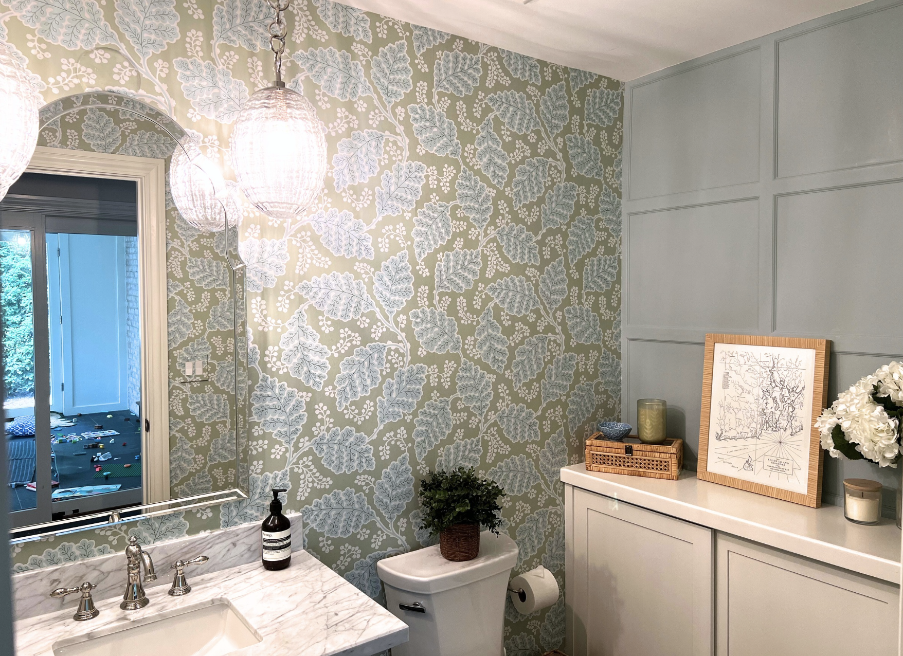 A bathroom with floral wallpaper, a mirror, a sink with a marble countertop, a soap dispenser, potted plant, a cabinet with decorative items on top, a framed art piece, and a wall with painted wainscotting above a custom cabinet.