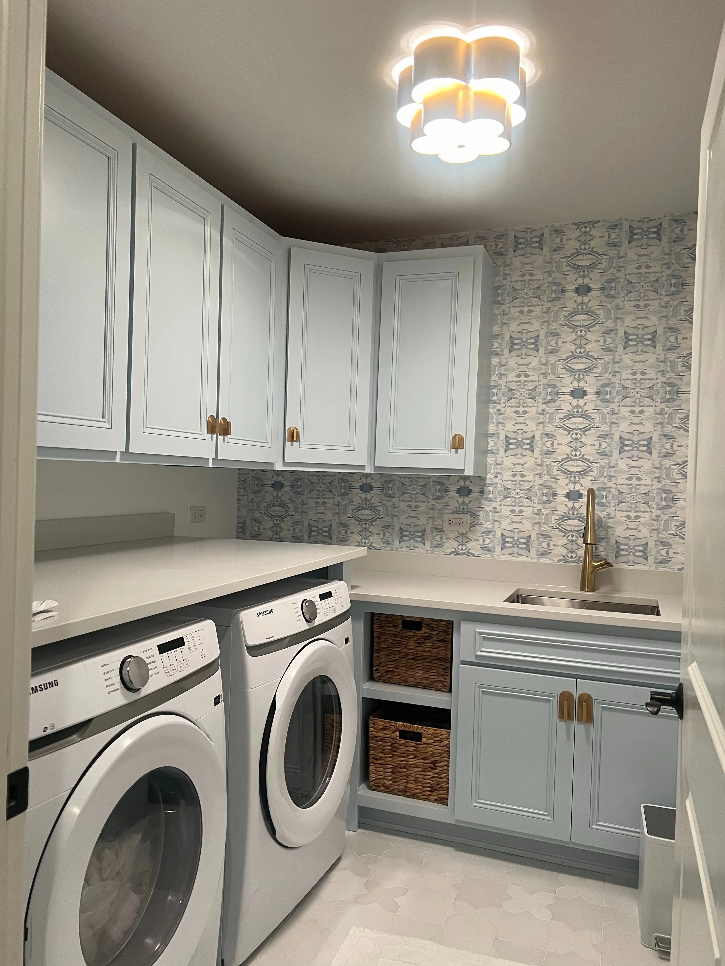 Laundry room with white cabinets, a washing machine, a dryer, a small sink with a gold faucet, and woven baskets in open shelves. Blue and white patterned wallpaper on one wall, white tile flooring, and a modern ceiling light fixture.