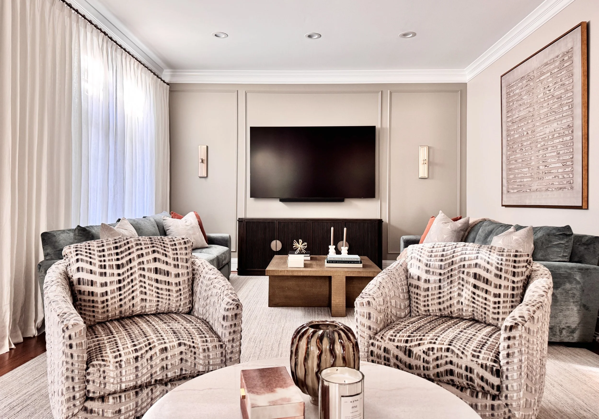 Modern living room with two patterned armchairs, two gray sofas, a wooden coffee table, a wall-mounted flat-screen TV, and decorative items.