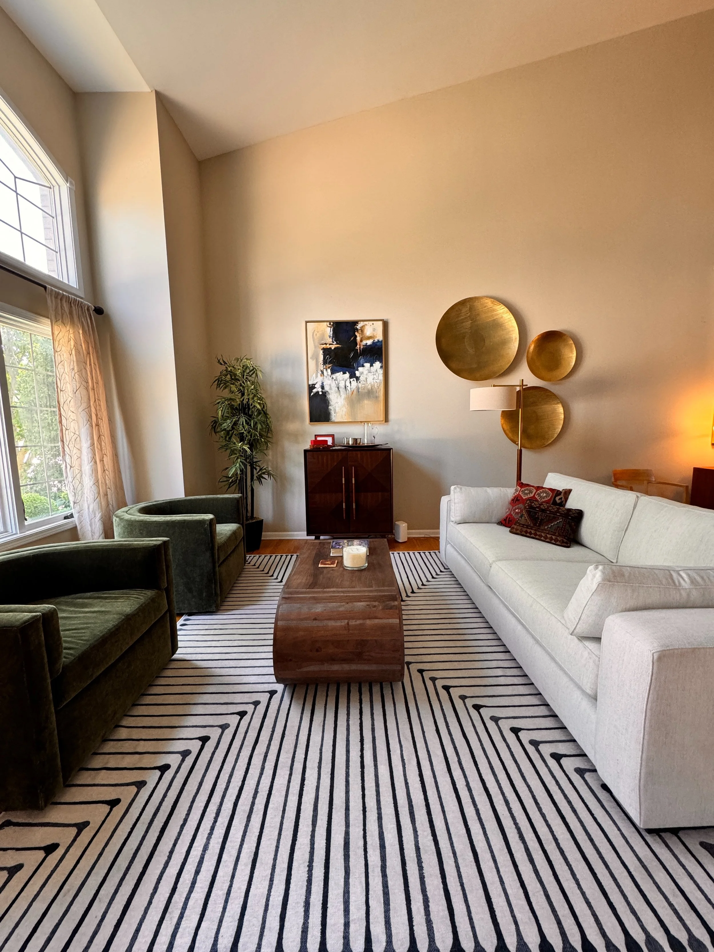 Living room with a white sofa, two green armchairs, a wooden center table, striped black and white rug, and modern wall decor including four golden circular wall art pieces. There is a large window with curtains on the left, a small cabinet with artw