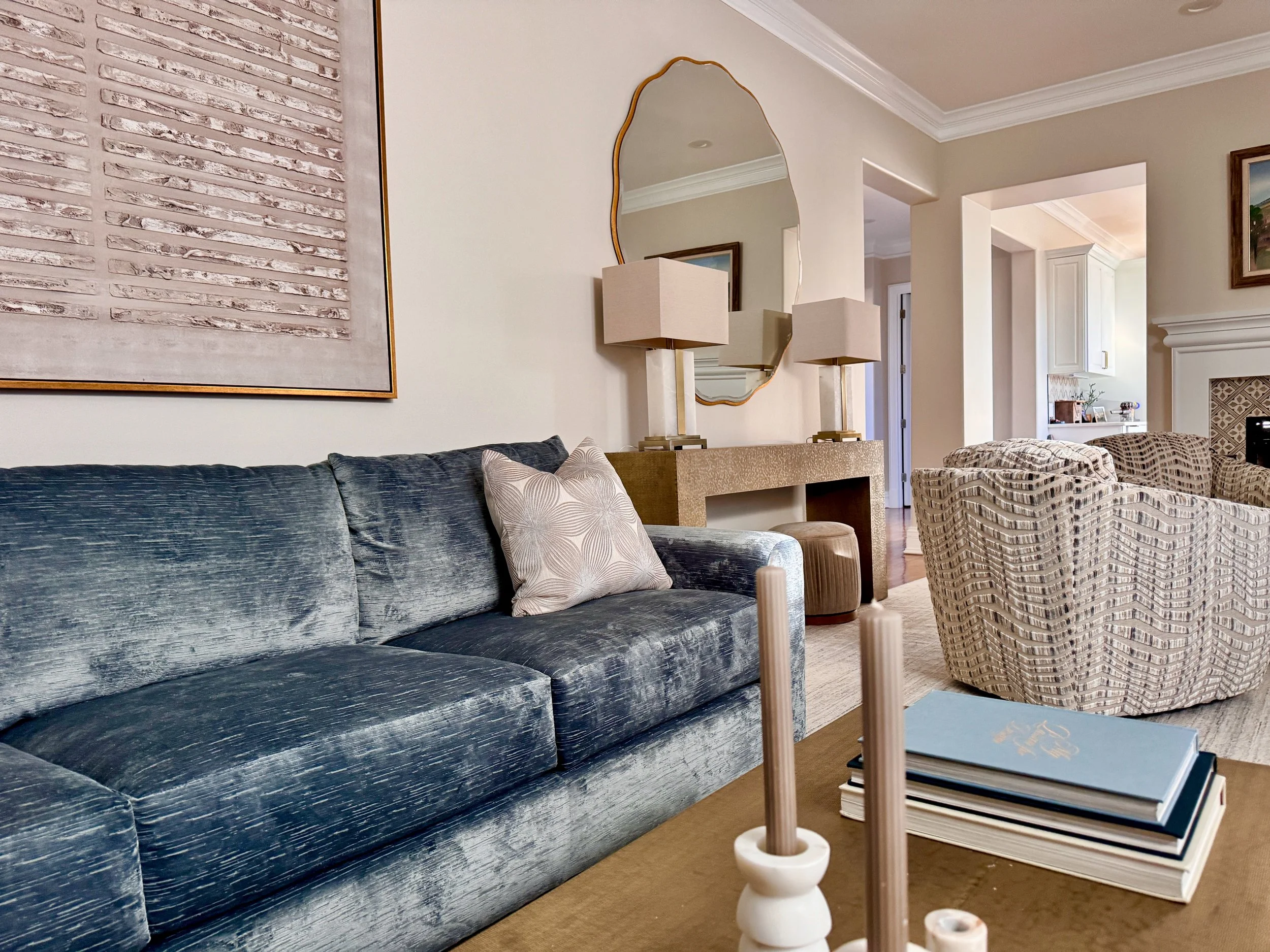 A living room featuring a distressed navy velvet sofa with a light-colored pillow, beige textured armchairs, a gold console table with lamps, a large mirror, and wall art, with a view into a kitchen and hallway.