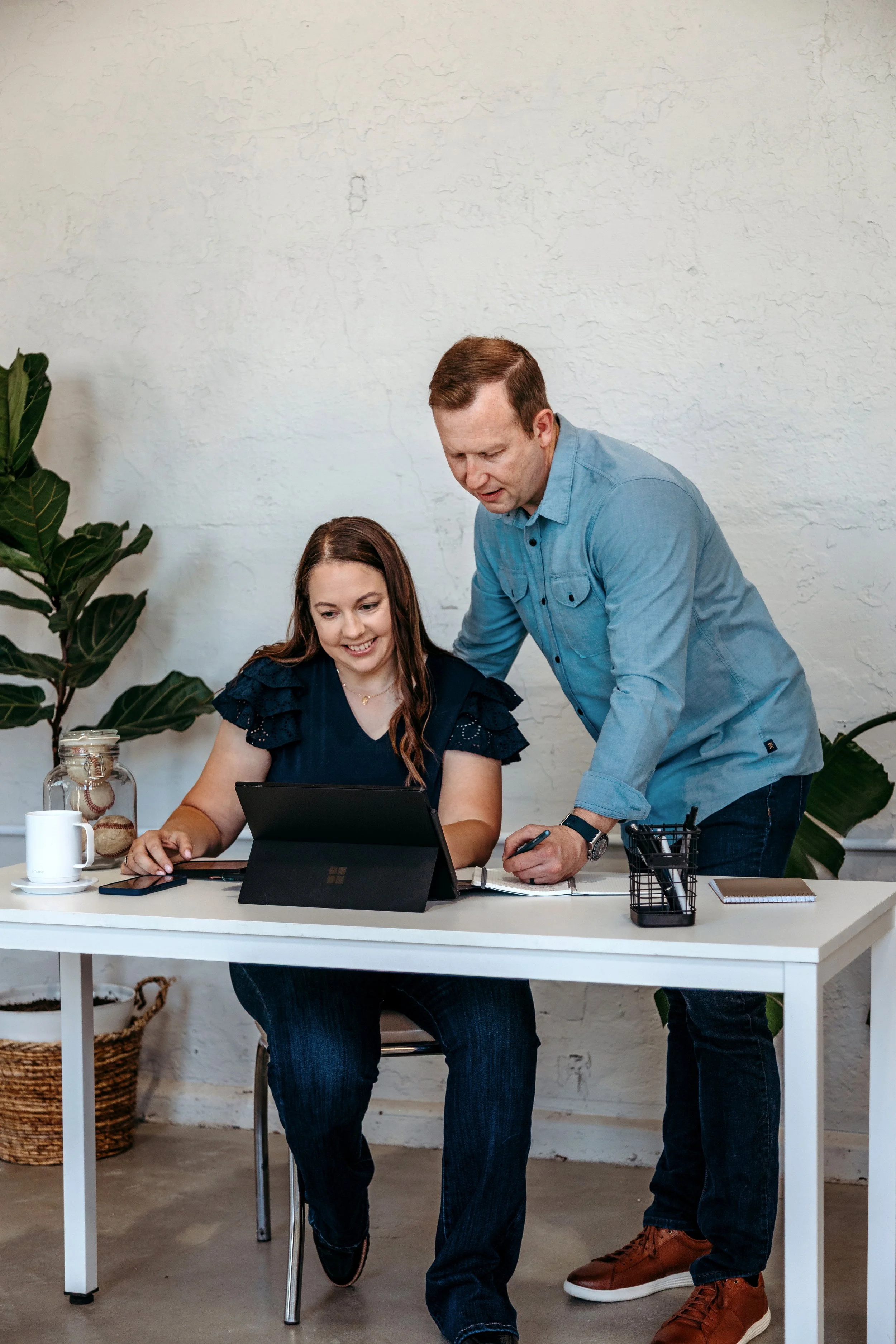 A man and a woman working together at a desk, looking at a tablet. The woman is seated, smiling, and the man is standing, pointing at the tablet. There are plants and office supplies on the desk.