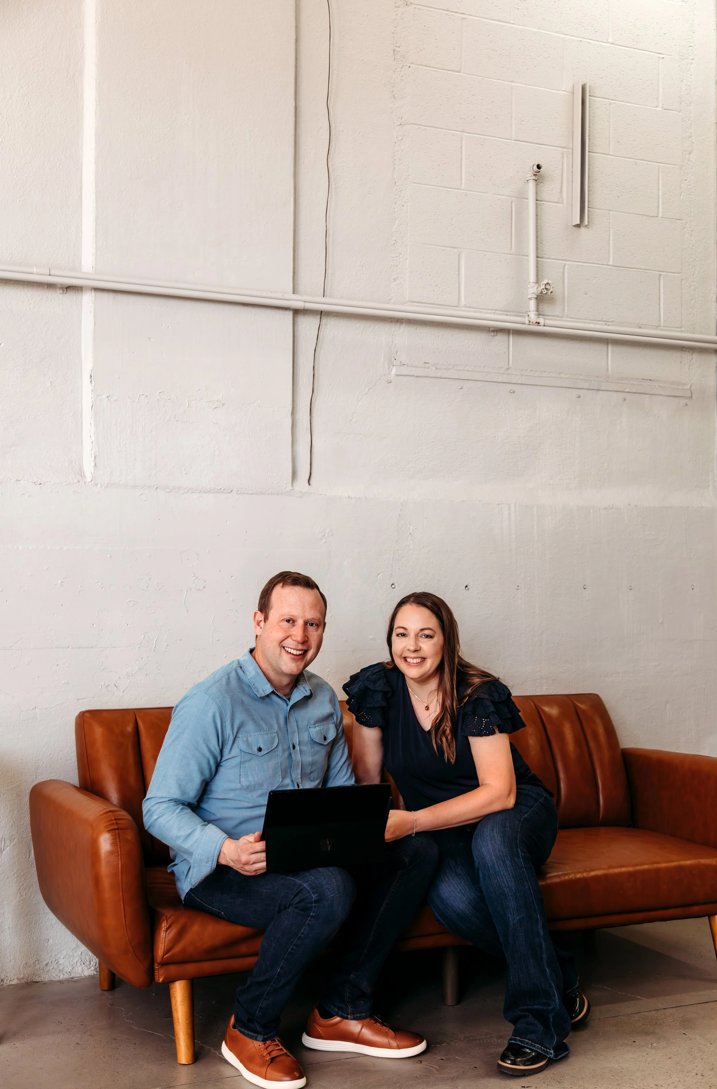 A man and woman sitting on a brown leather couch, smiling, with a laptop.