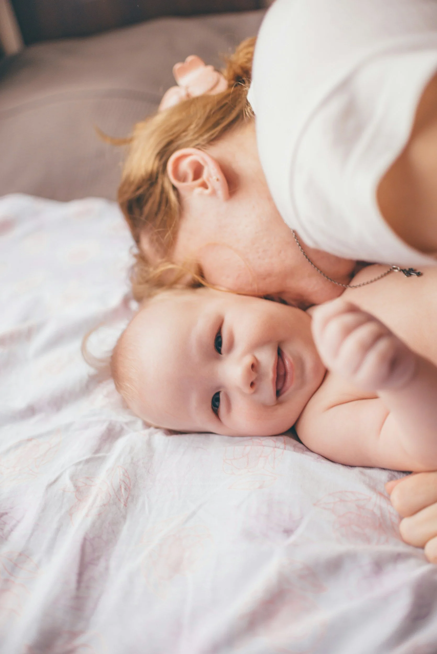 A woman with red hair and a black necklace lying on her side, resting her head on a baby with light-colored skin, dark eyes, and a smile, on a bed with light bedding.