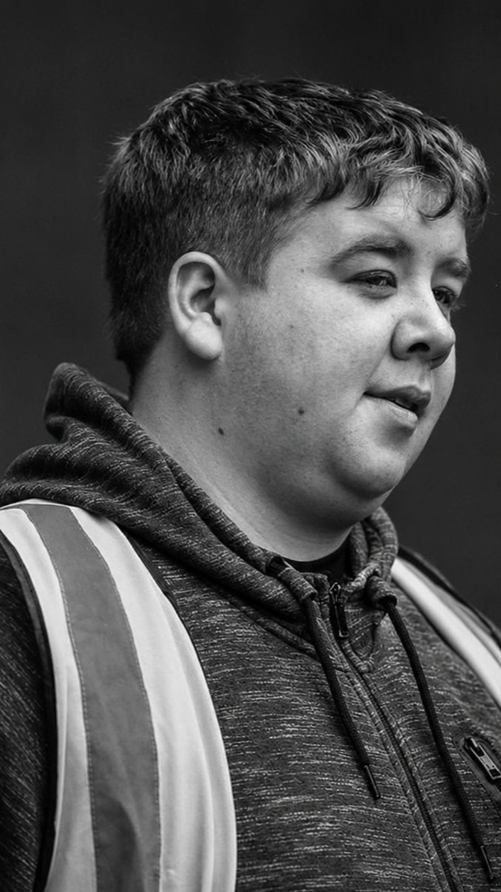 A young man with short hair wearing a dark hoodie and a reflective safety vest, smiling slightly and looking to the side.