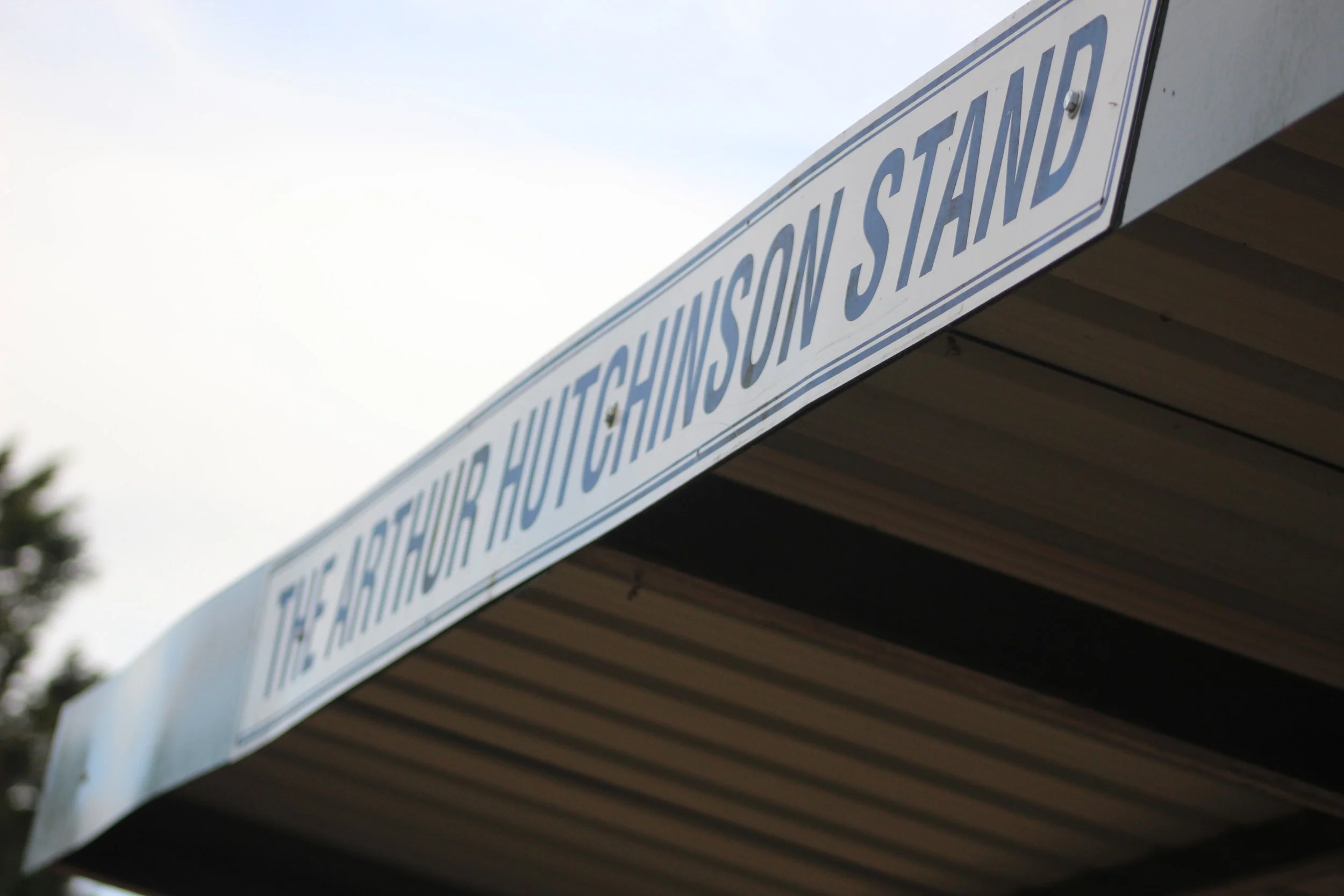 Close-up of a white sign with blue text reading 'THE ARTHUR HUTCHINSON STAND' attached to a corrugated roof on top of a football stand.