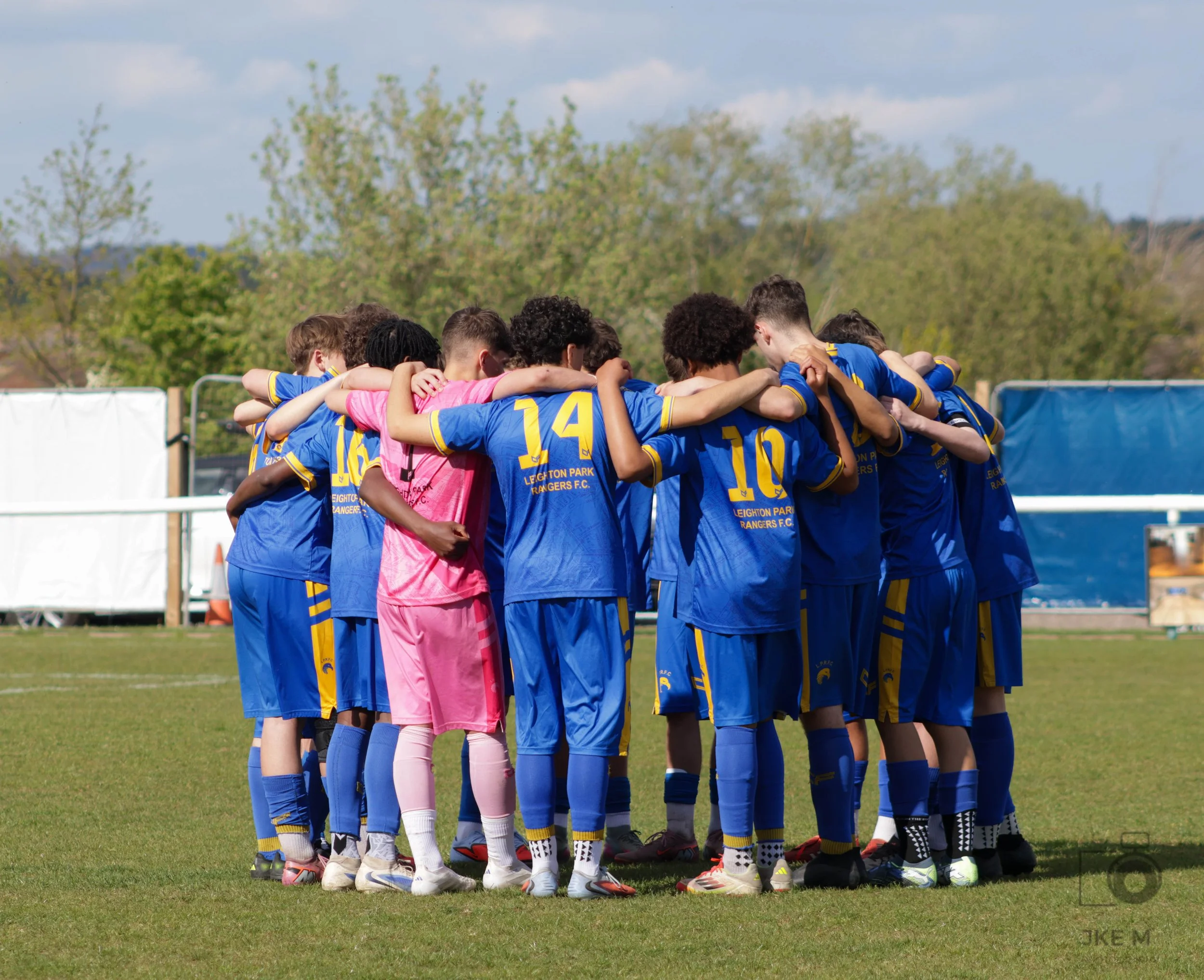 A group of footballers standing in a huddle on a grass field, wearing blue with the goalkeeper in pink, with trees and a cloudy sky in the background.