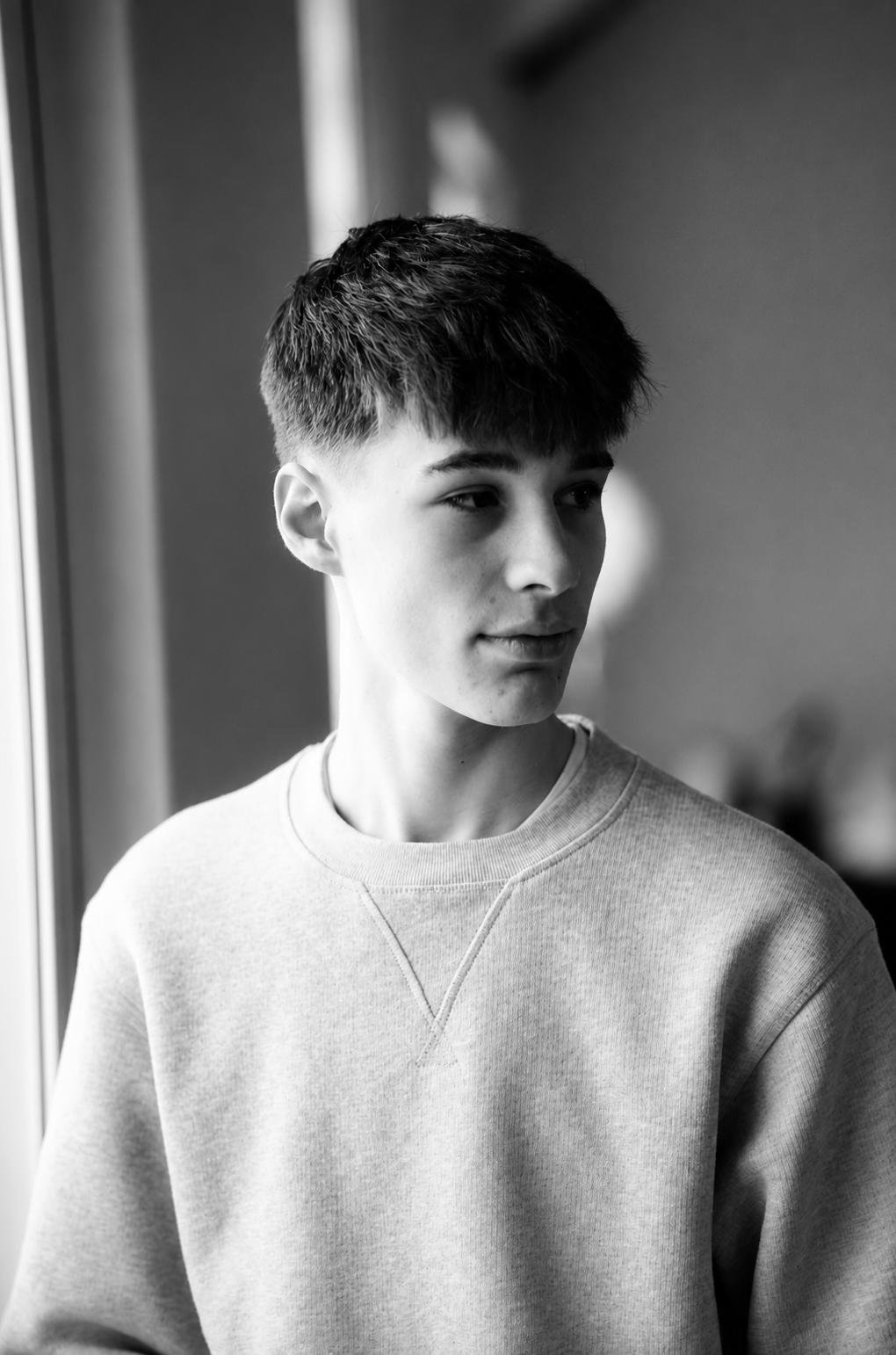 A black and white photo of a young man with short hair looking to the side, wearing a casual sweatshirt, indoors near a window.