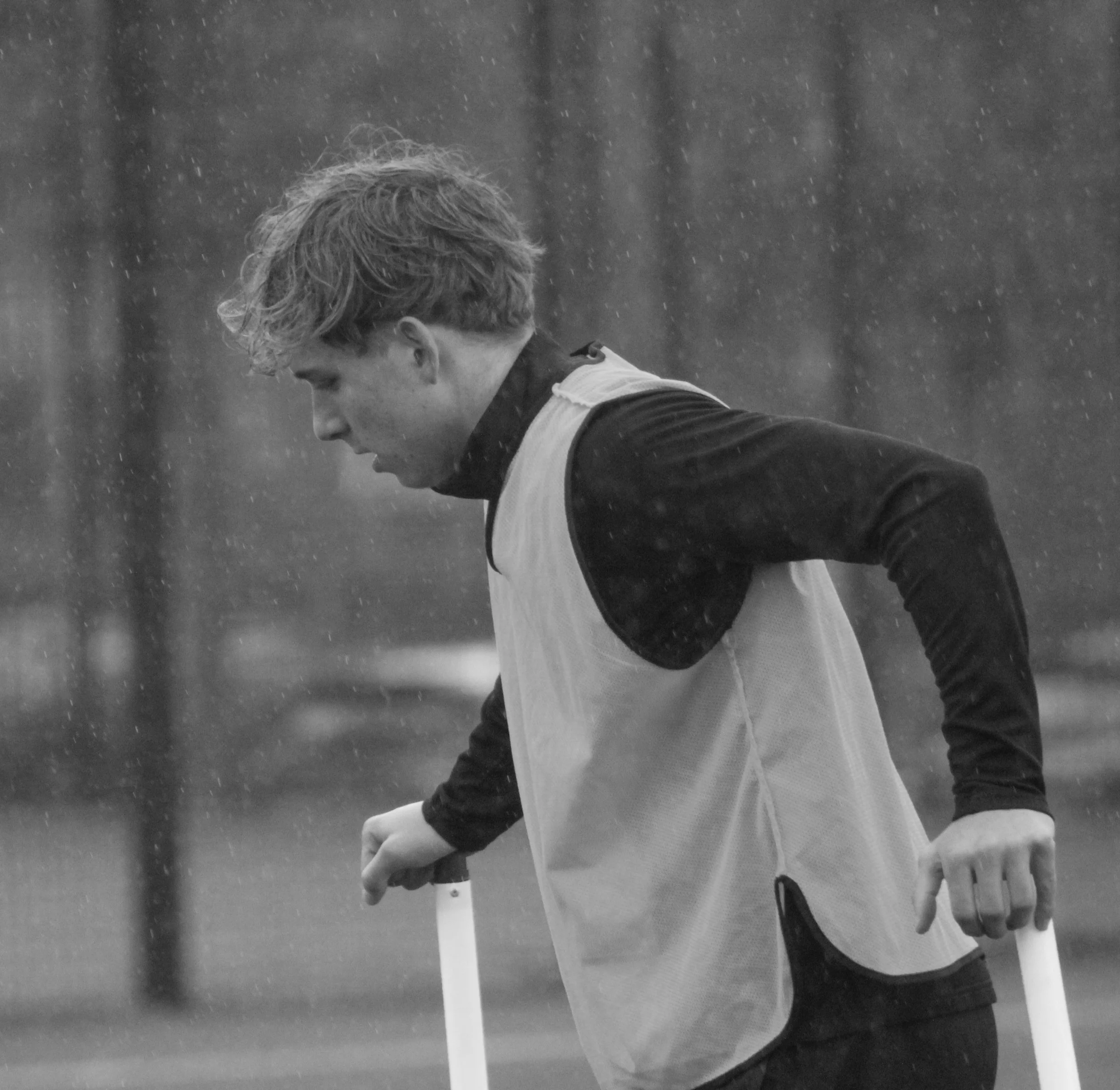 A young man playing football  in the rain looking down. He is wearing a dark shirt and a light vest, with rain falling around him.