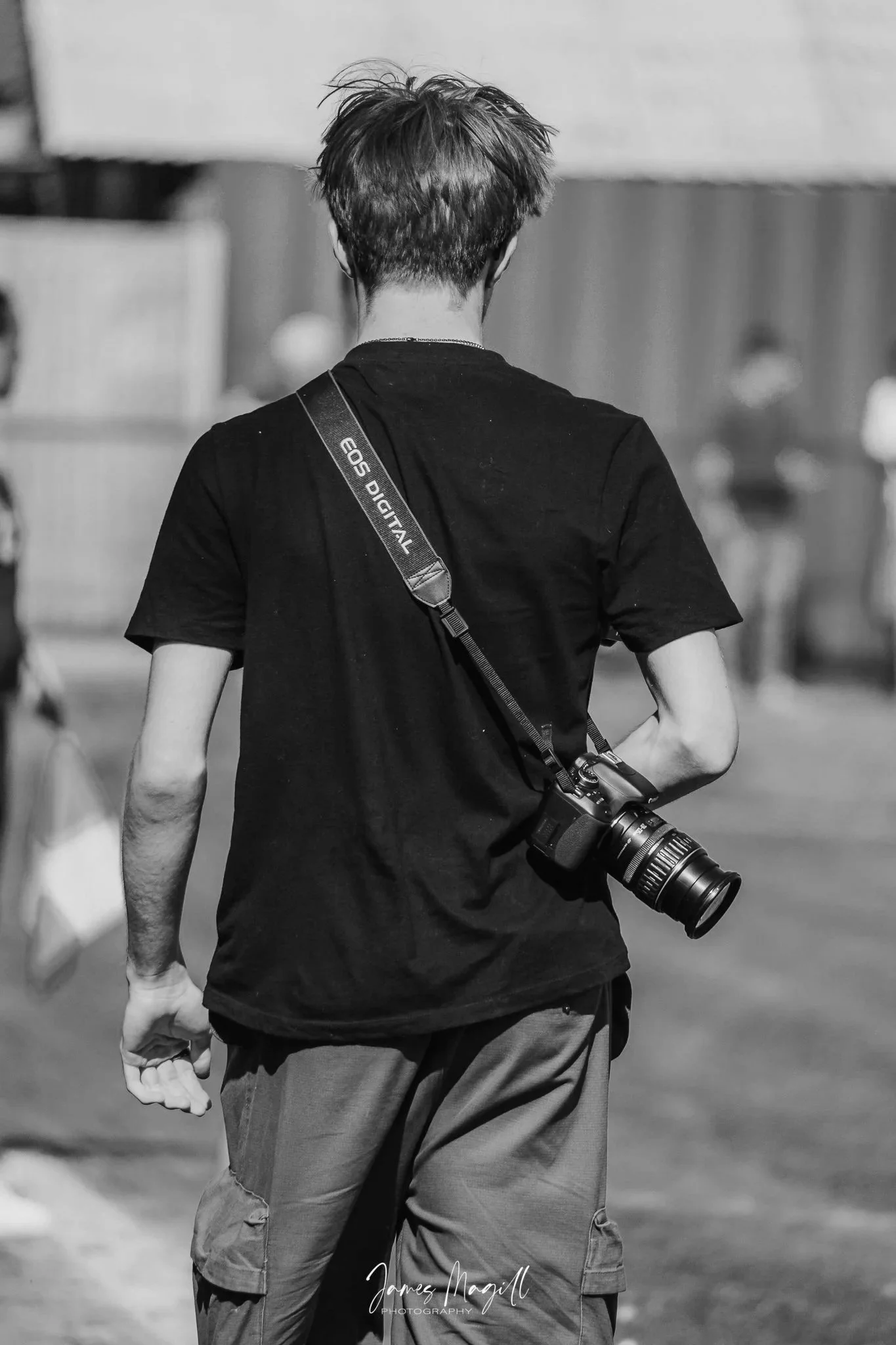 A person with messy hair wearing a black t-shirt and khaki shorts with a camera hanging from their shoulder, walking outside.