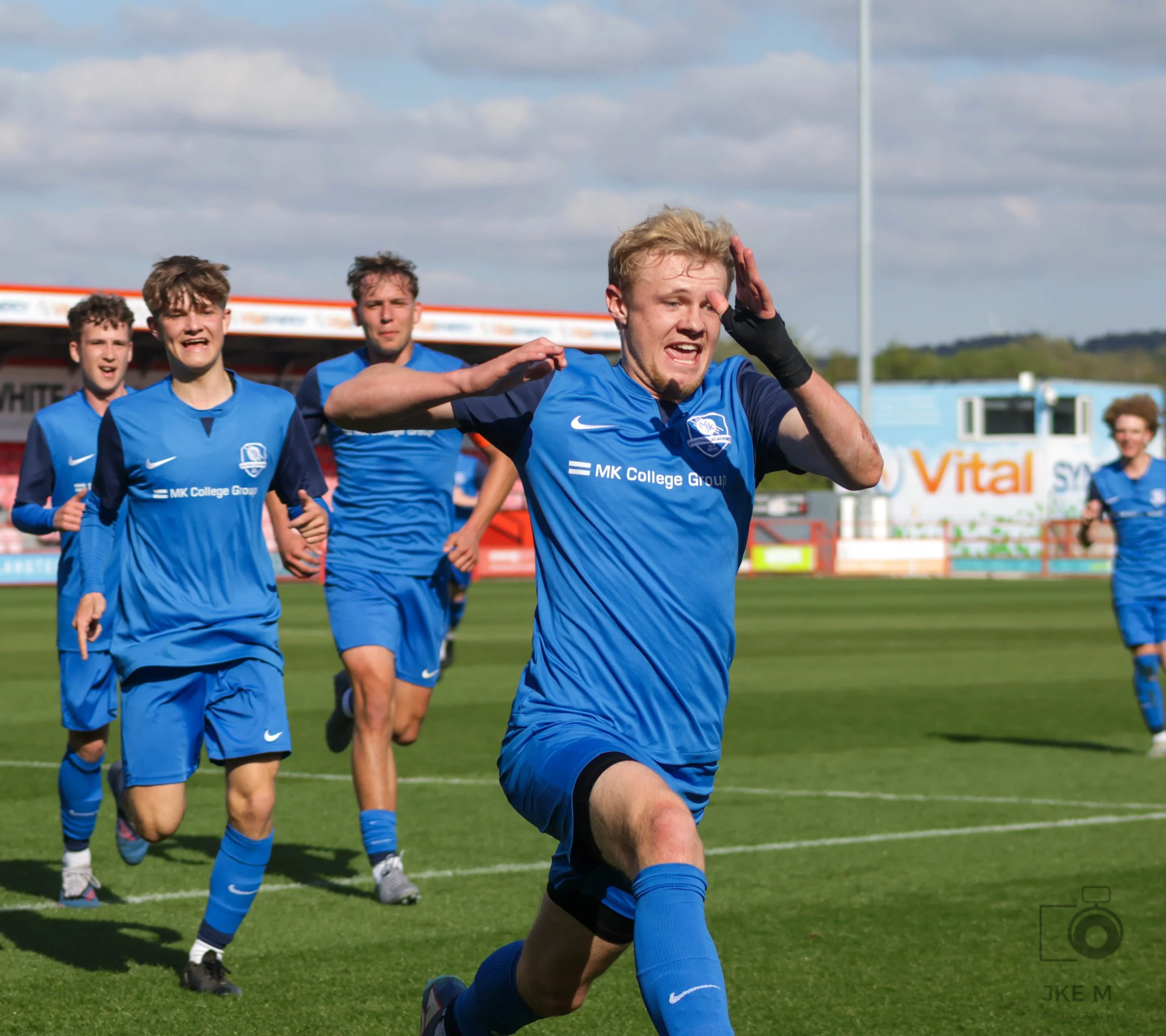 A group of young male soccer players in blue jerseys running on a soccer field during daytime; the player in front appears to be celebrating with his hand on his forehead.