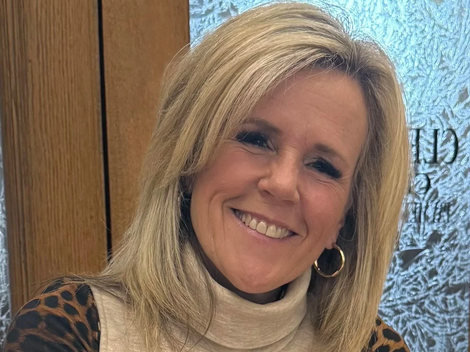 A smiling blonde woman with hoop earrings, wearing a light-colored turtleneck and a leopard print jacket. She is standing indoors near a textured, possibly frosted glass window and a wooden wall.