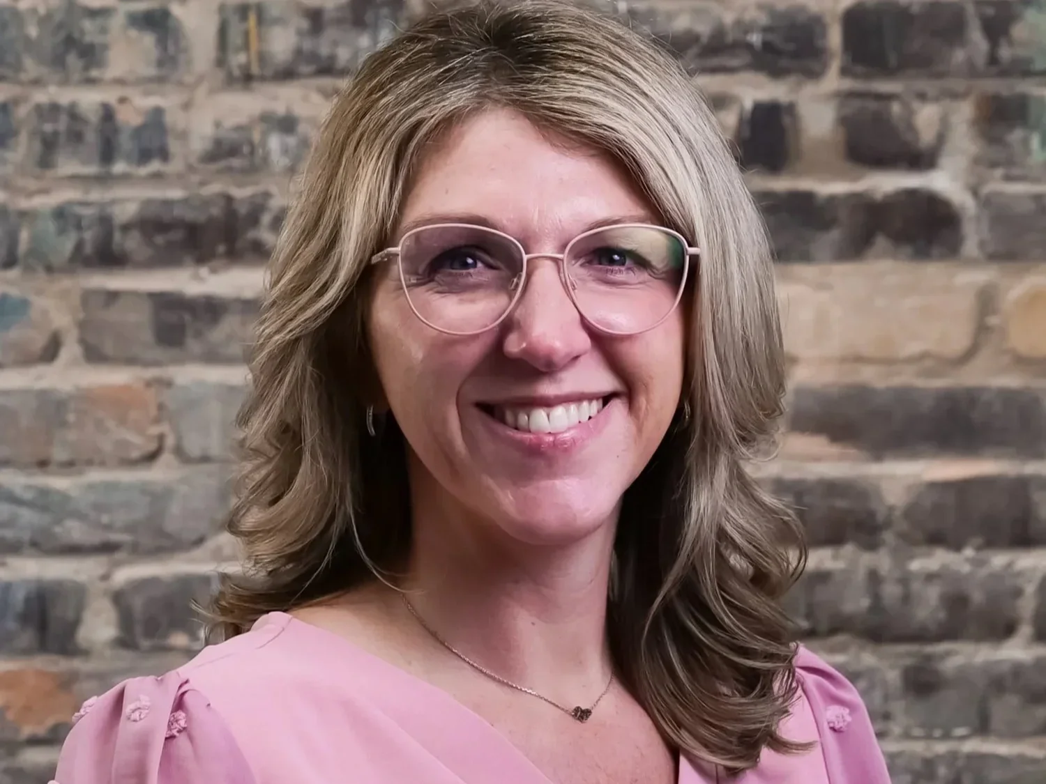 A woman with shoulder-length wavy hair, wearing glasses and a pink blouse, smiling in front of a brick wall.