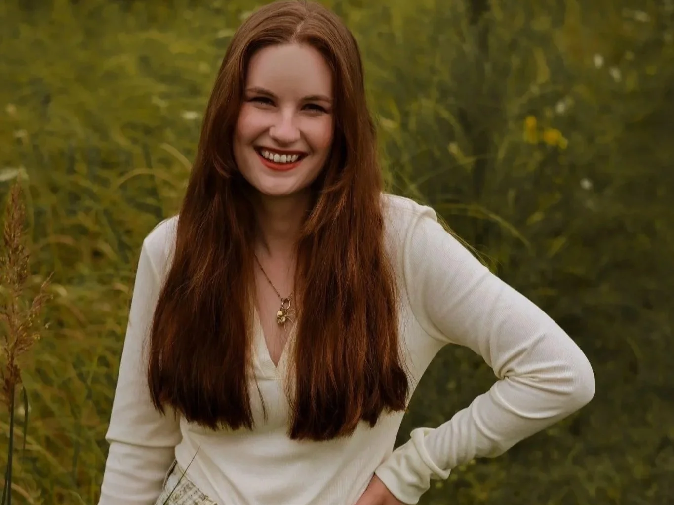 A young woman with long red hair smiling outdoors in a natural setting with green foliage in the background.