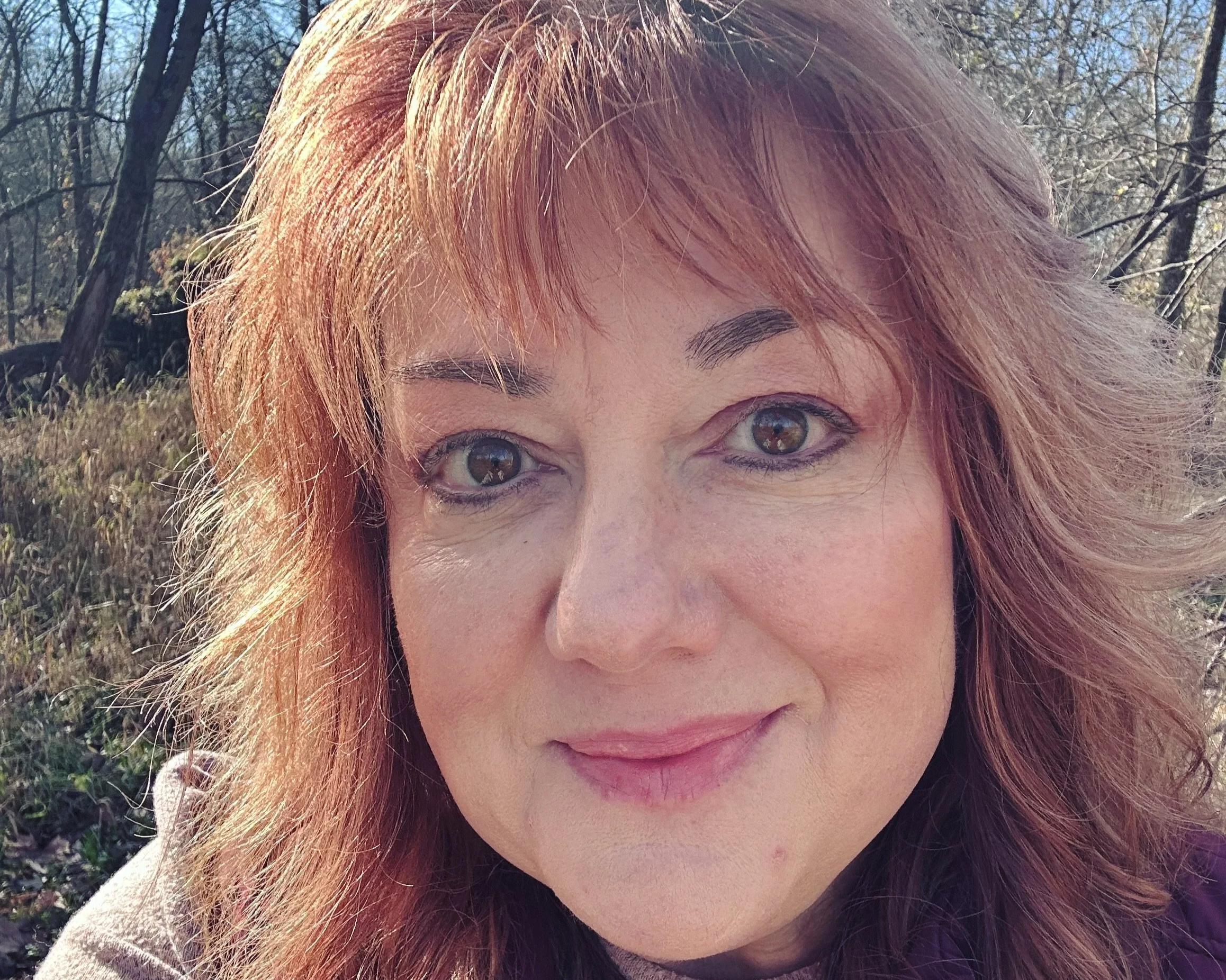 Close-up of a smiling woman with wavy reddish-brown hair outdoors in a wooded area.