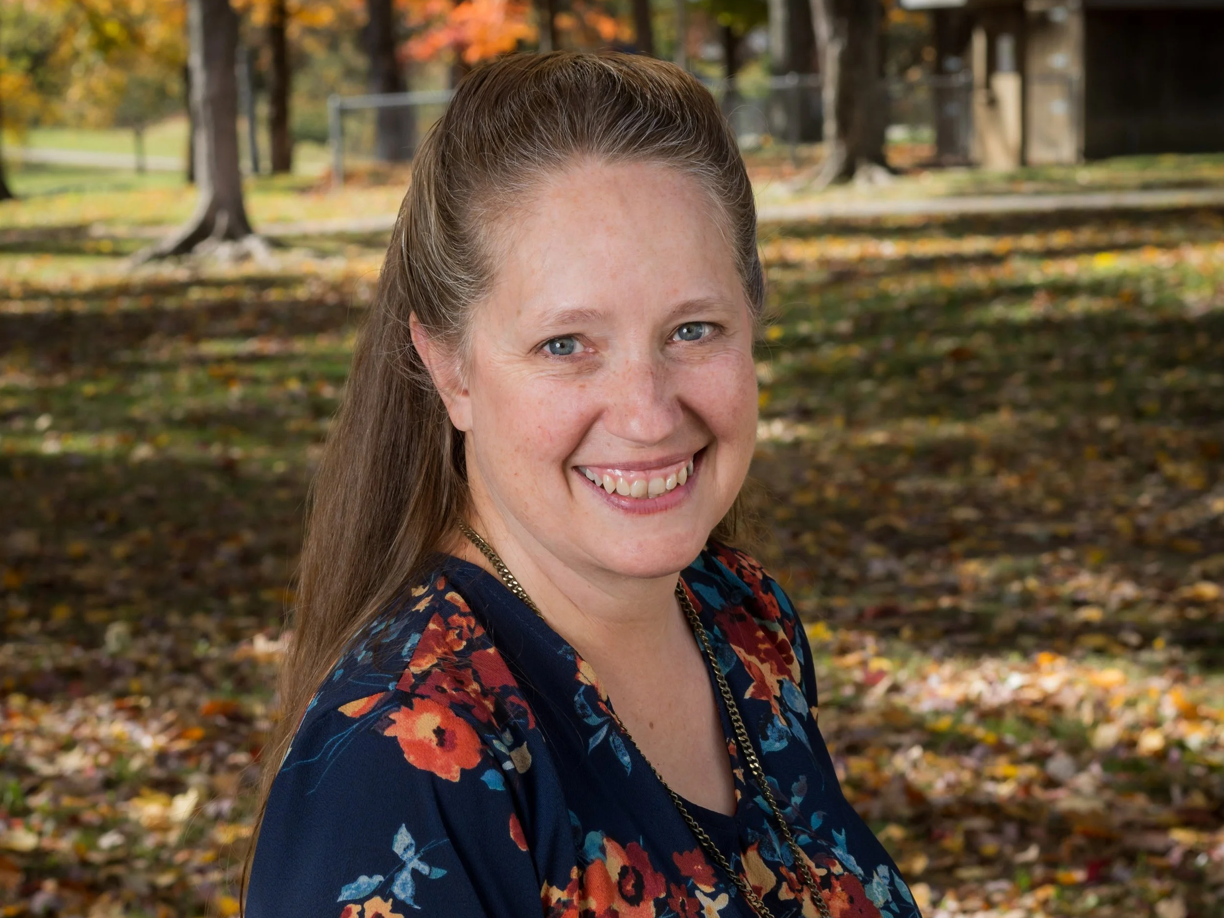 A woman smiling outdoors in a park during fall, with trees and fallen leaves in the background.