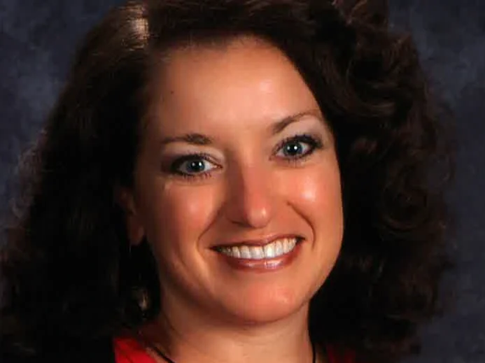 Close-up portrait of a woman with dark curly hair and blue eyes, smiling, wearing a red top, against a dark background.
