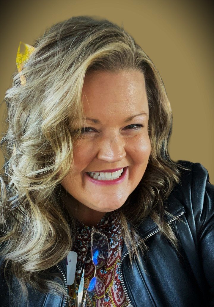 A woman with long, wavy gray hair, wearing a bright red dress, speaking at a podium with a microphone in a conference or event setting.